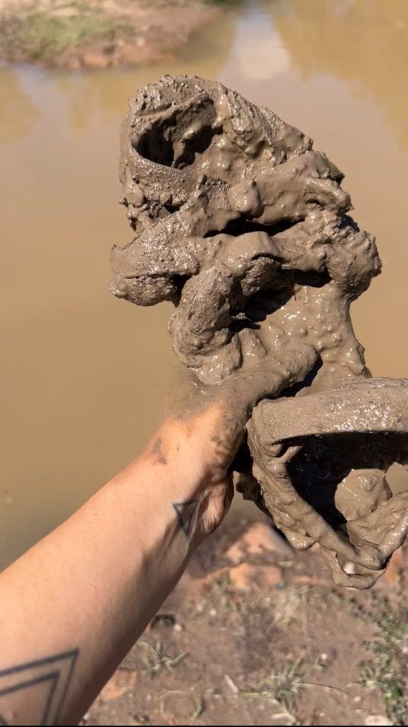A hand holding a Teva Sandals covered in Mud in front of a muddy quarry.