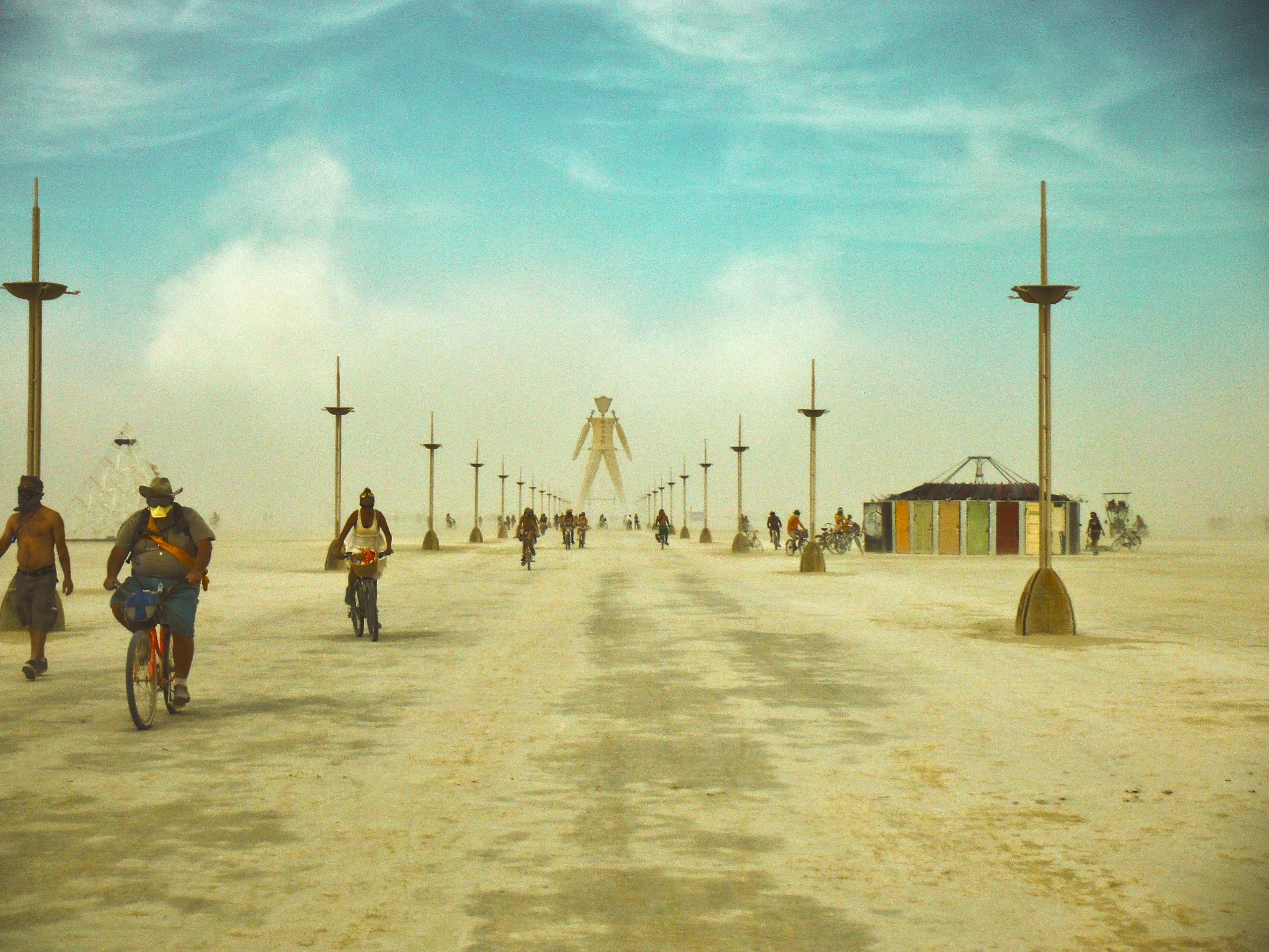 People riding bicycles along a dusty path in the desert of Black Rock City aka Burning Man leading to the wooden Man structure. In the distance there are some colorful art installations.