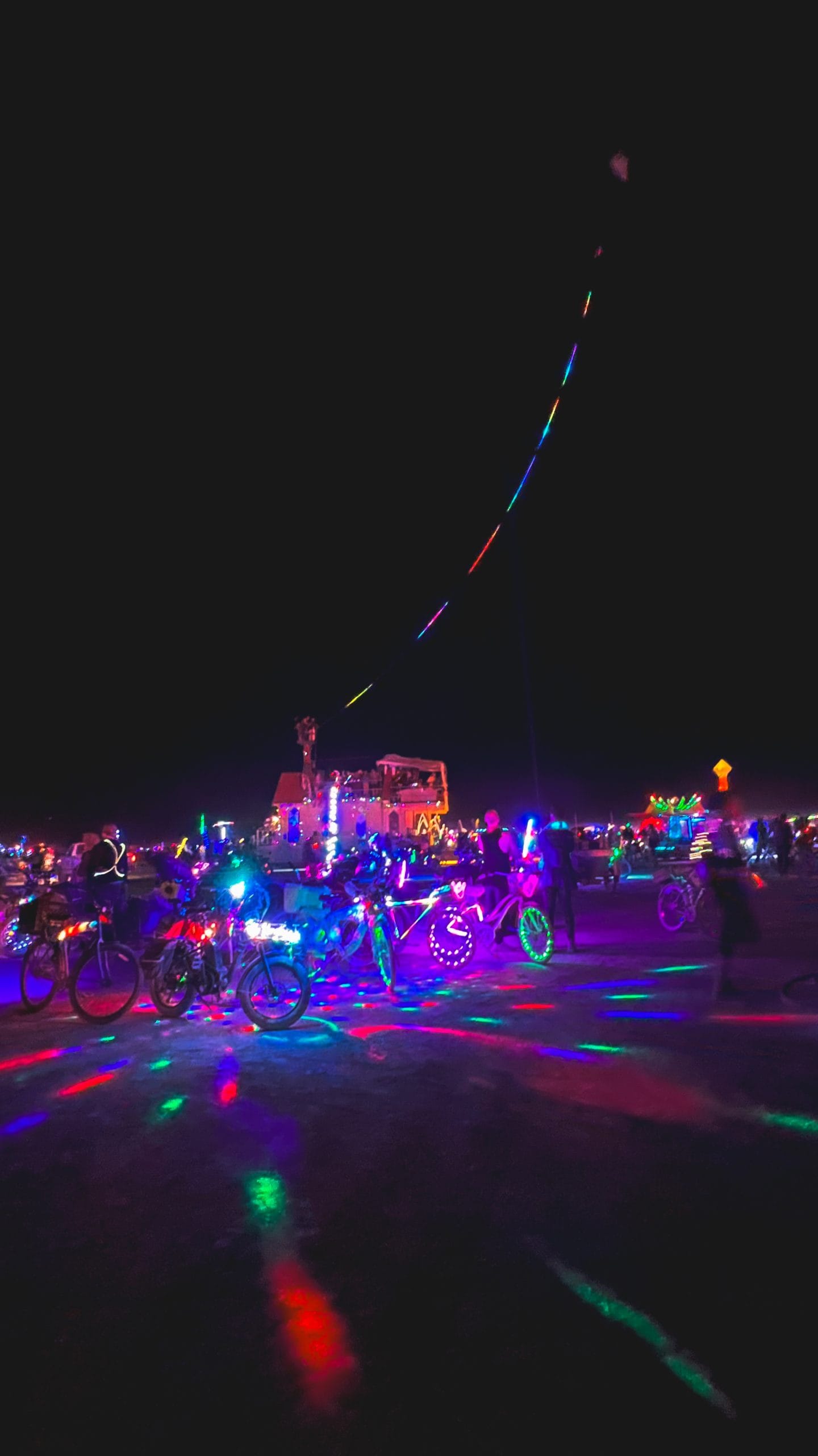 Colorfully lit bicycles gathered on the playa at night during Burning Man, with neon lights reflecting on the ground and art cars glowing in the background.