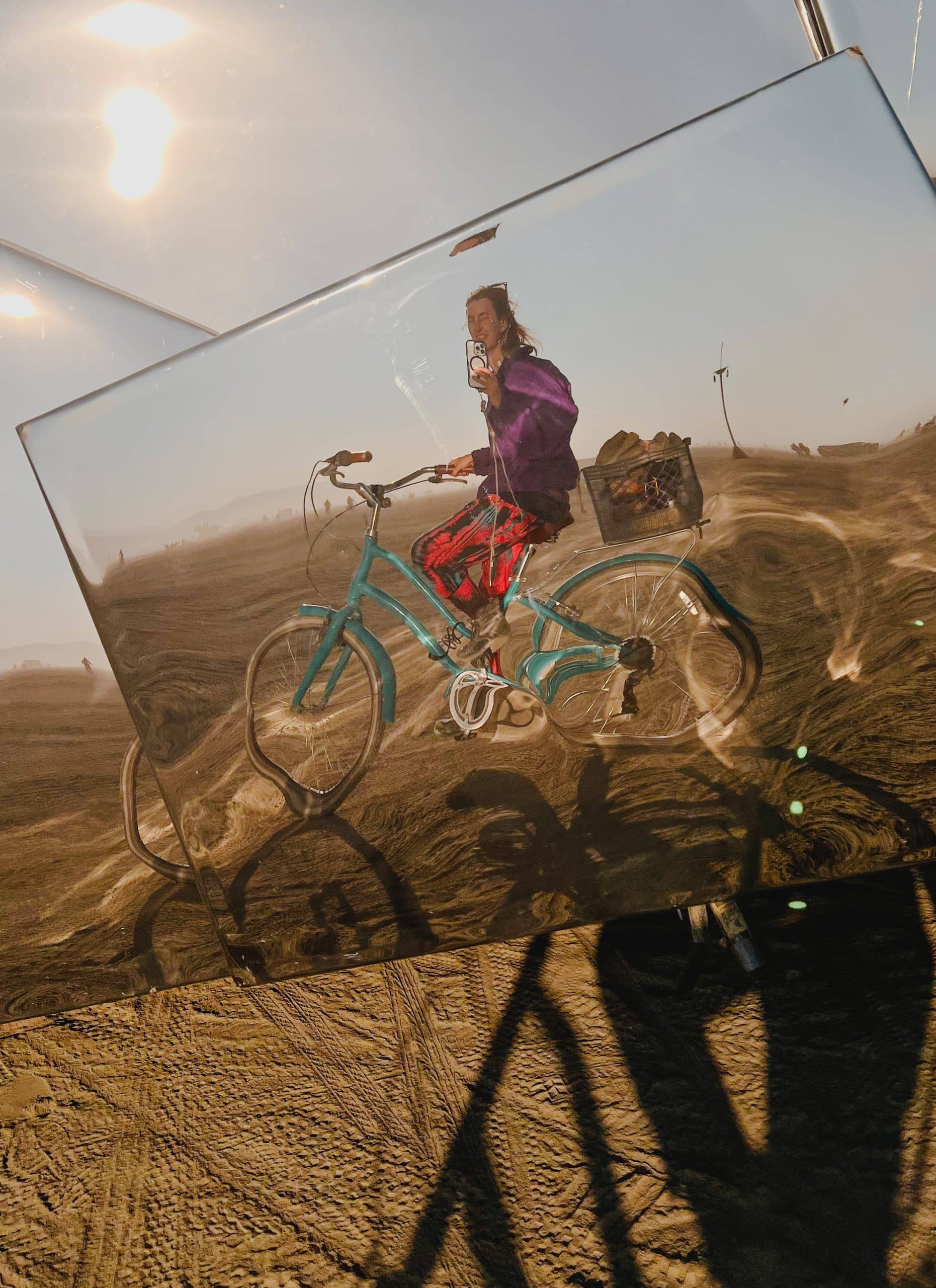 Blogger Tasha Jacobs taking a photo with her phone of her reflection from a shiny reflective moving art car while riding a bike at Burning Man. The reflection of the bike is warped.