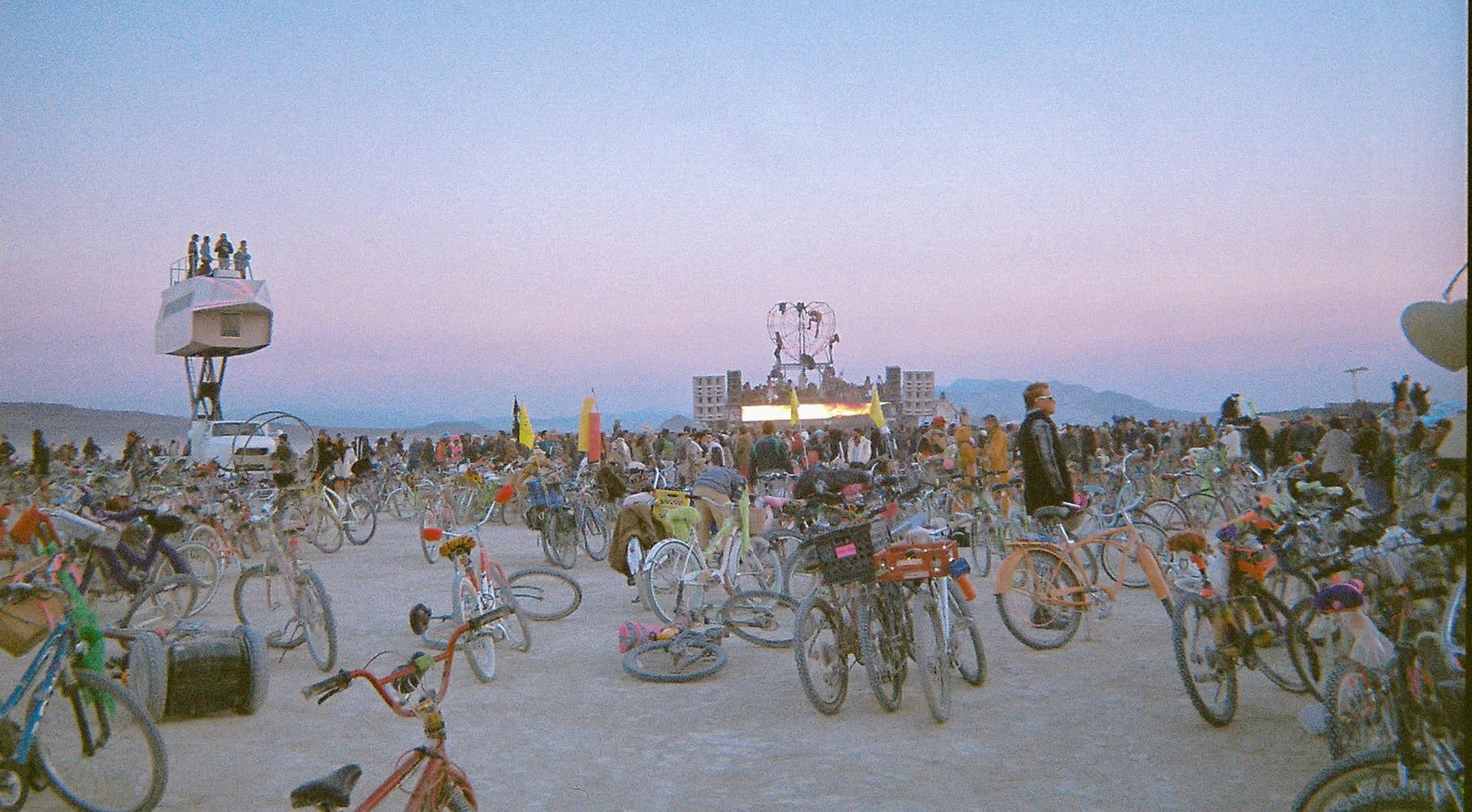 Hundreds of decorated Burning Man bikes and Burners gathered on the playa near art installations at sunset in Black Rock City.