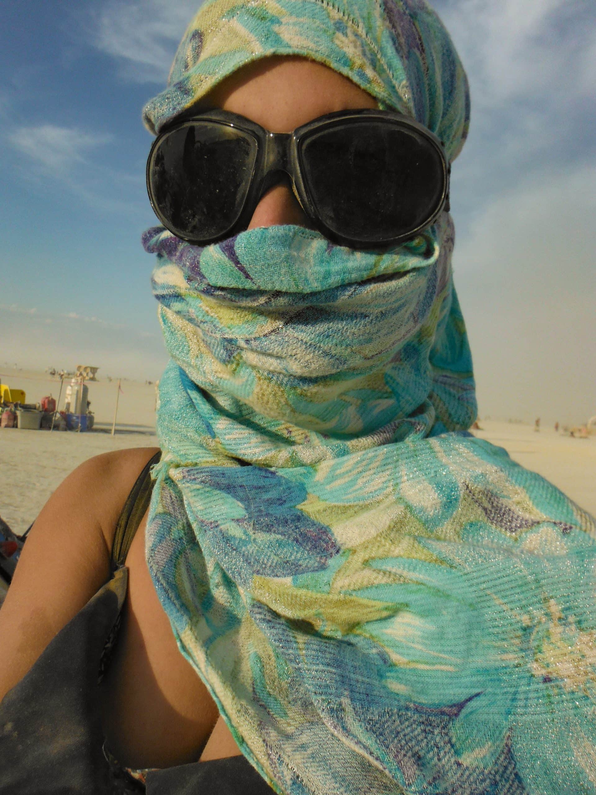 Tasha at Burning Man wearing a blue floral scarf wrapped around her head, mouth, and neck only revealing her goggles. Slight dust storm and blues skies in the background.