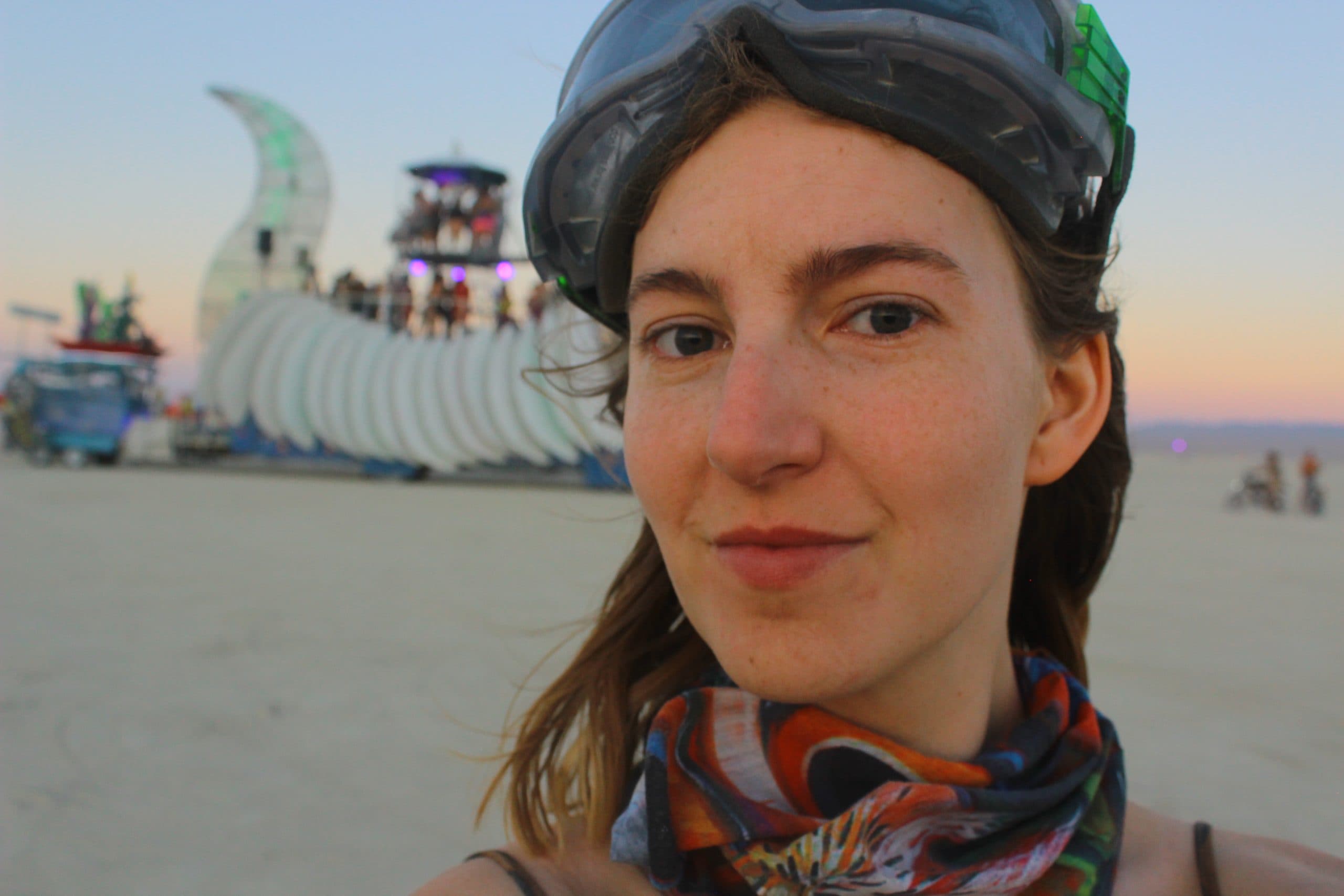 Tasha looking at the camera at Burning Man with dust goggles on her forehead and a wrap around scarf around her neck. In the distance there is an art car and sunset.