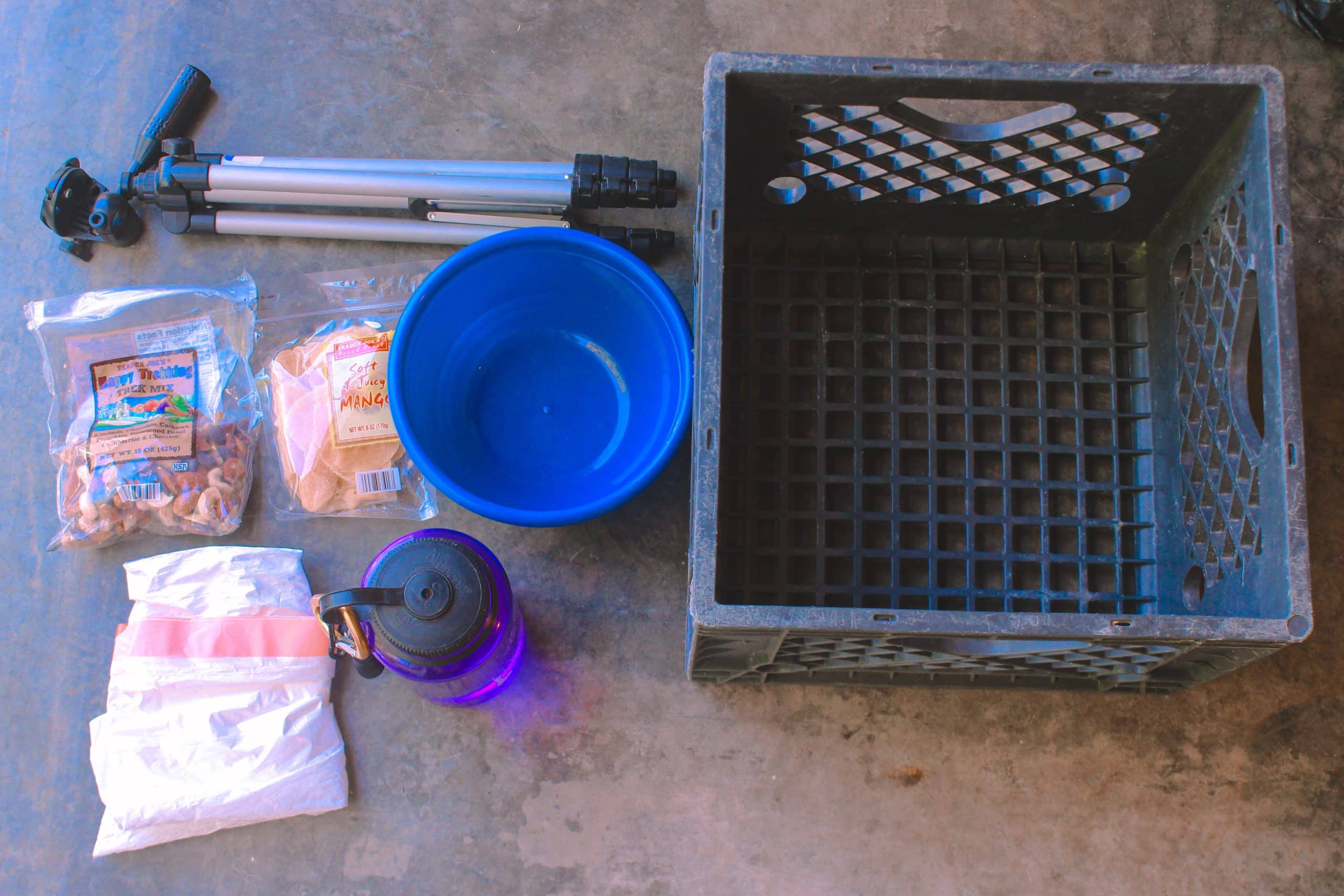 Milk crate and items like water bottle, bowl, tripod, garbage bag, and snacks laid out on the floor ready to pack for Burning Man.