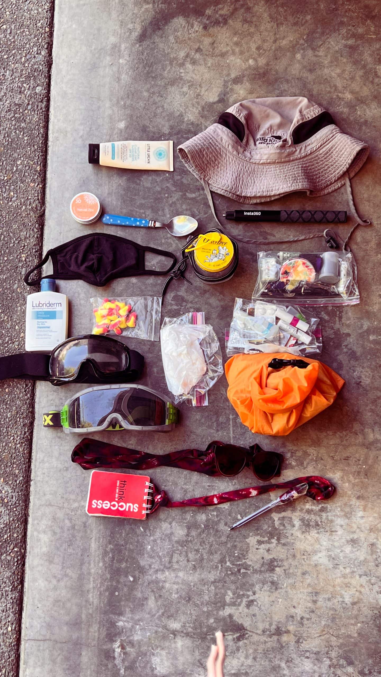 Many items laid out on the ground ready to be packed for Burning Man event including dust goggles, sunhat, lotion, portable cup, and sunscreen.