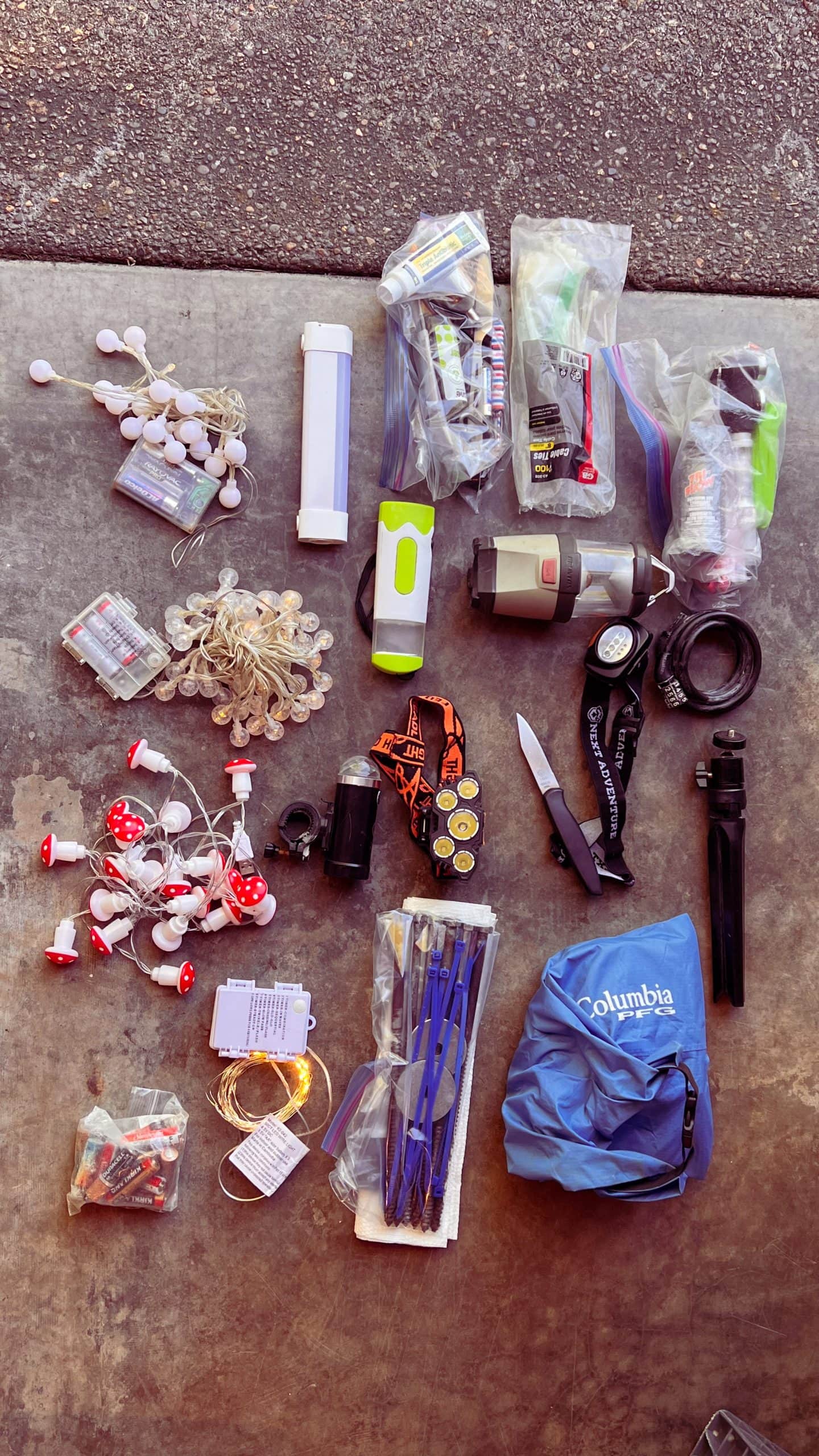 Several items laid out on the ground to pack for the festival Burning Man including bike lights, bike lock, lantern, dry bag, and tripod.