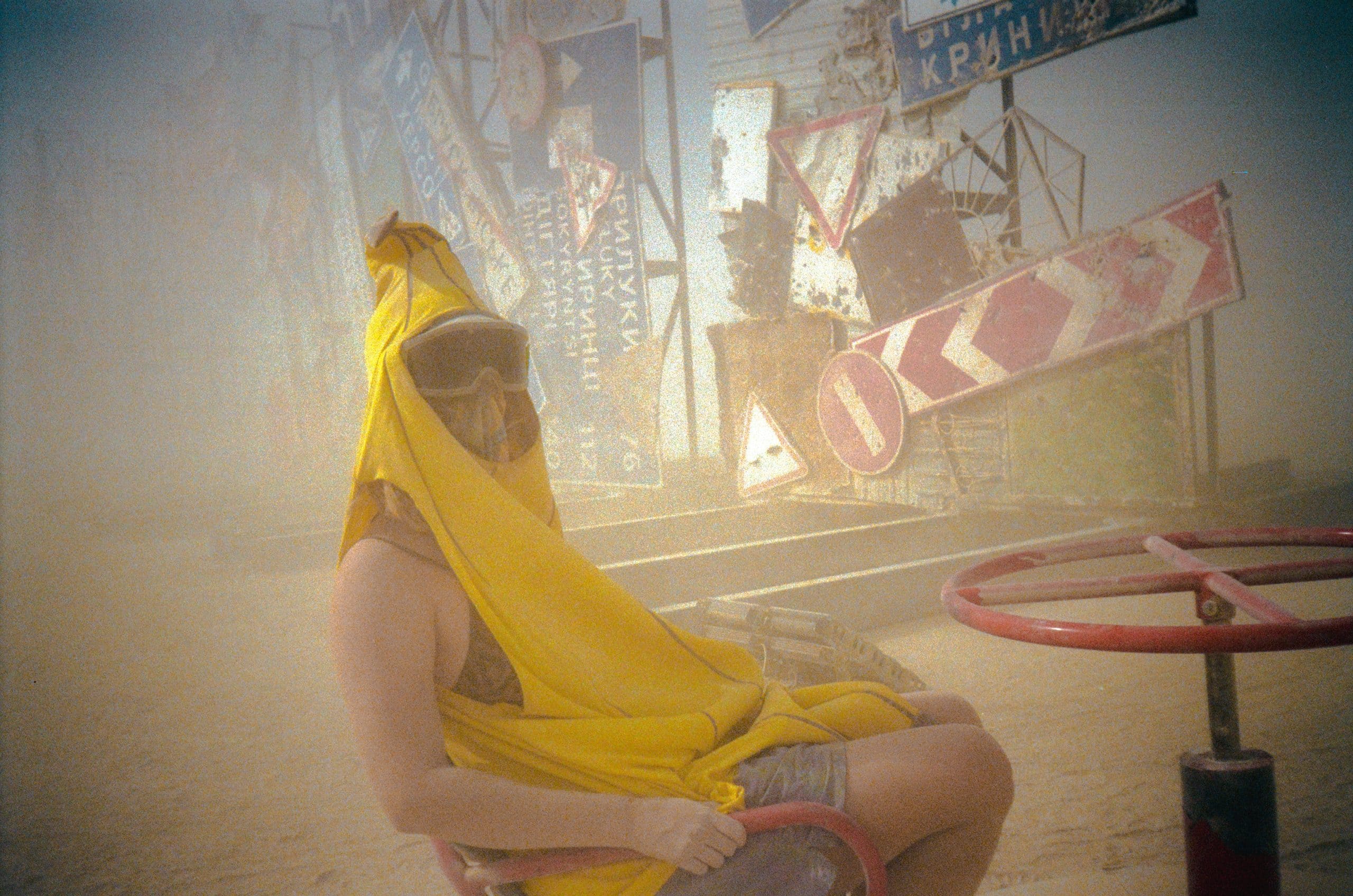 Tasha the travel blogger in a banana suit and dust goggles sitting down at Burning Man in a slight dust storm. The Ukrainian art instalation "I'm Fine" is barely seen in the distance in front of the dusty air.