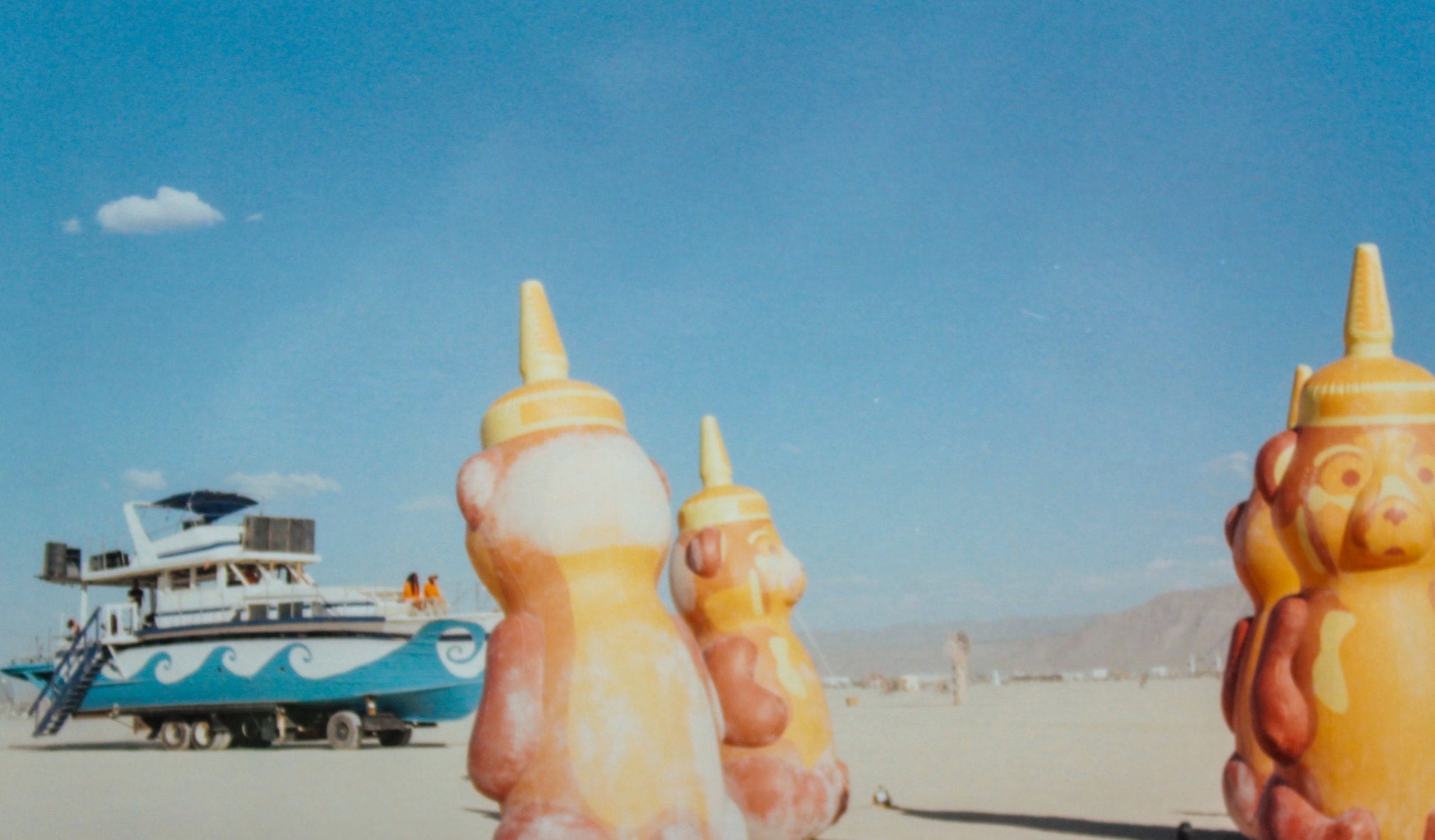 Art installation at Burning Man that resembles squeezable honey bear containers. Three giant bears in the desert with a boat art car in the distance.