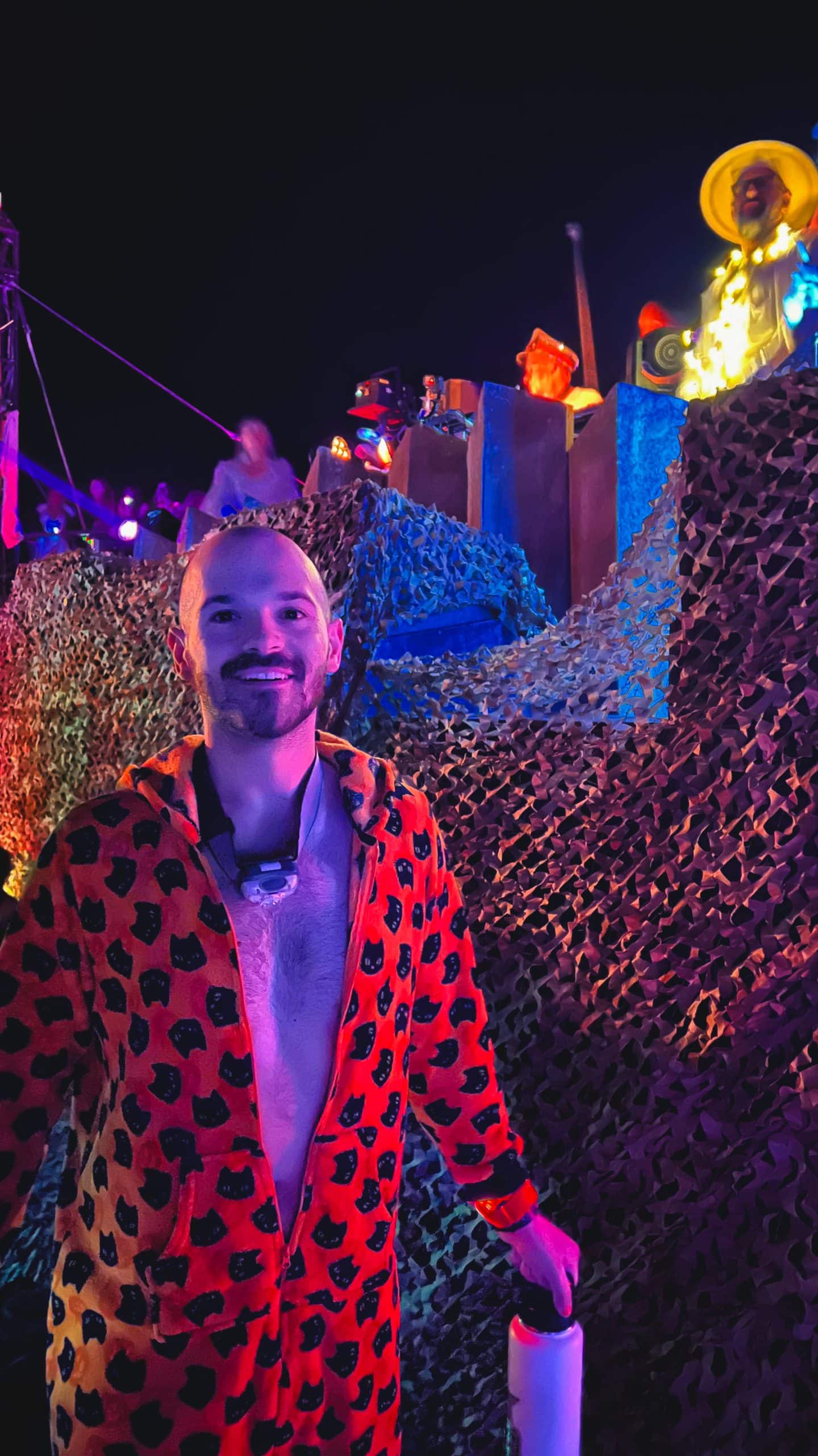 Man at Burning Man wearing a headlamp around his neck and a orange onesie with a sound stage in the background at Burning Man.
