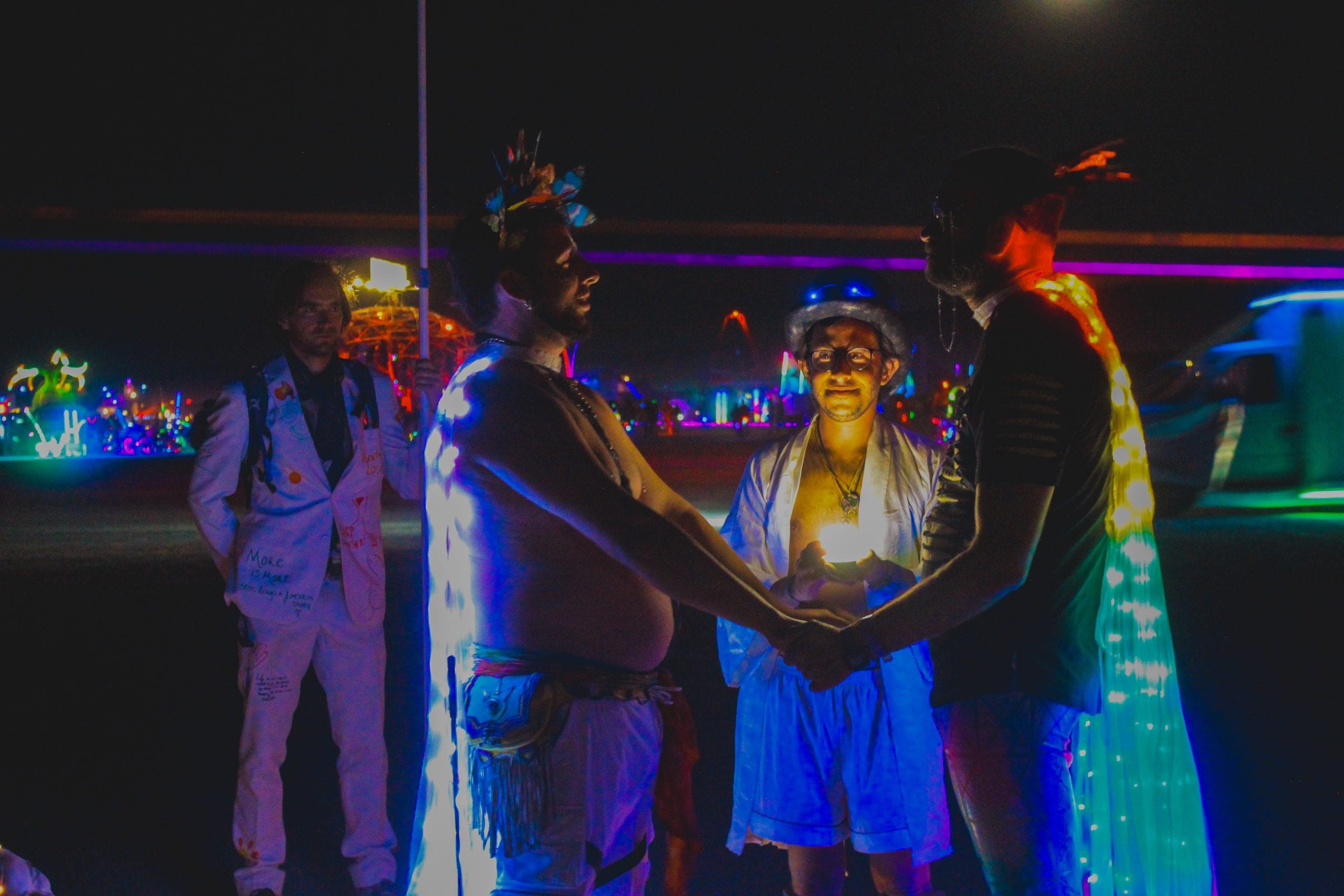 Two lovers getting married at Burning Man during night time. Their neon light capes are lit up vibrantly and in the distance there are laser lights making straight parallel lines. More lights in the background and on the officiant as well as lit up art cars. 