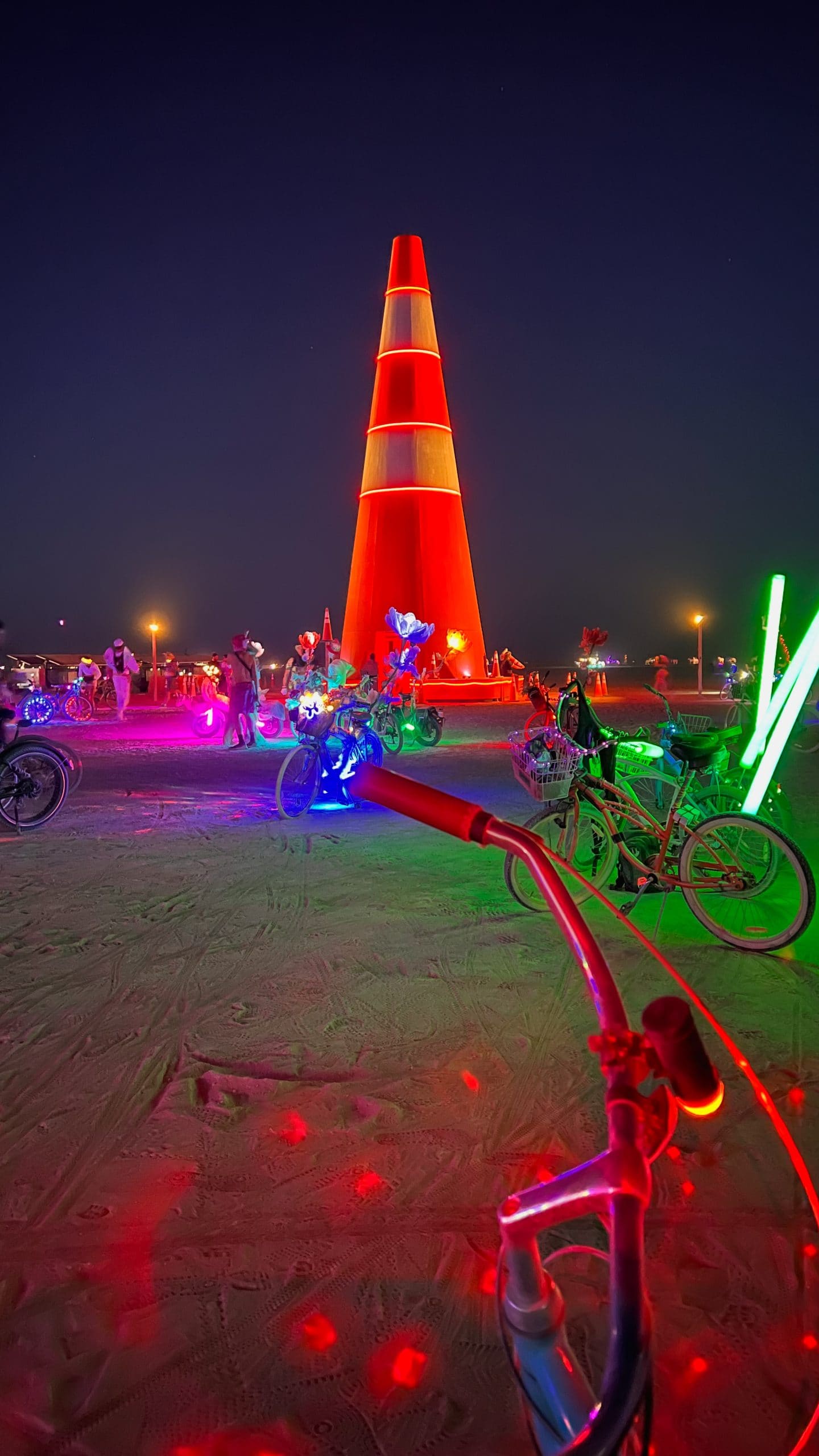 A Burning Man bike with a disco light illuminating the ground below. The giant traffic cone and other neon lights glow in the night time dark in the distance.