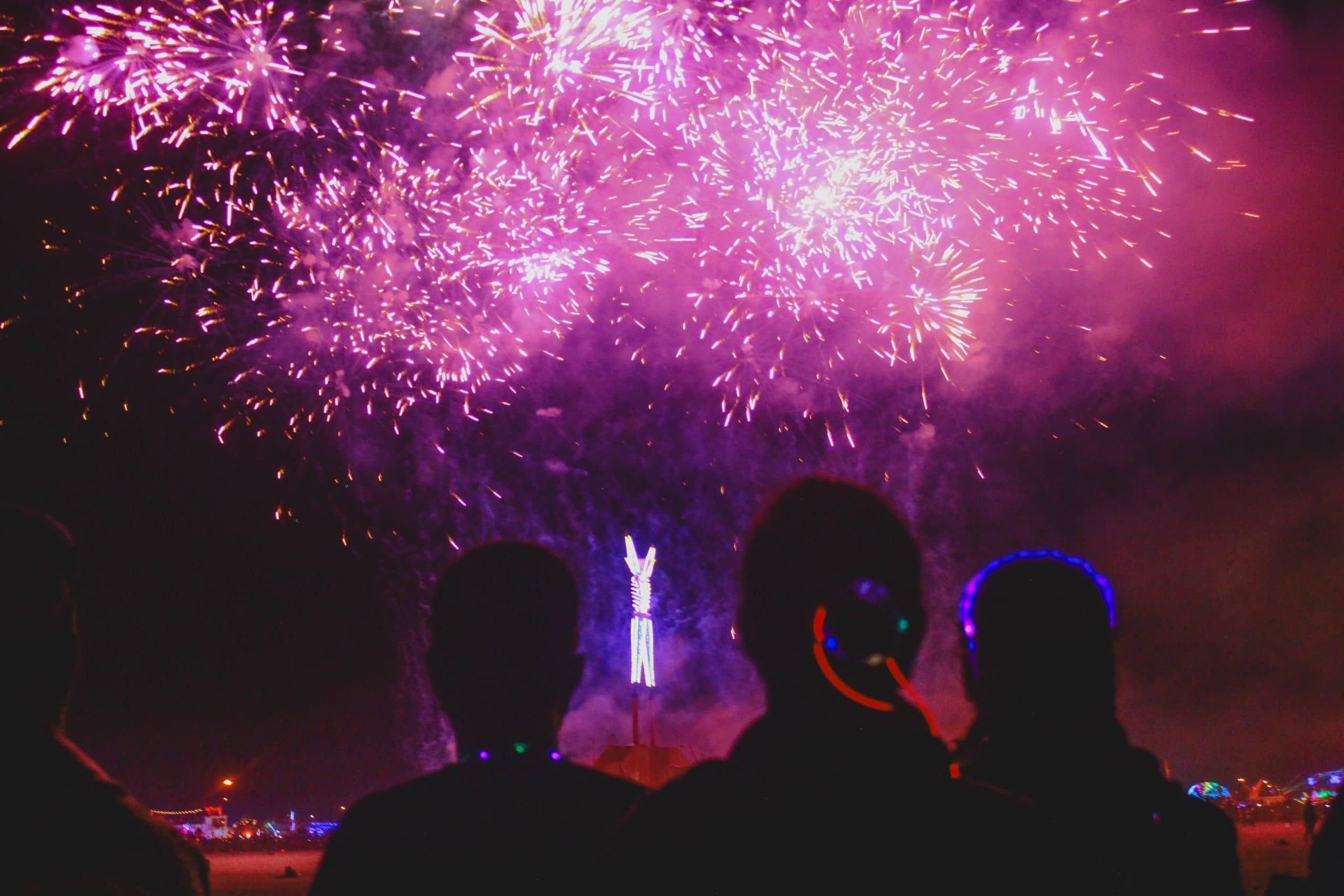 In the distance, the Burning Man structure lit up with purple lights and the sky full of exploding purple fireworks. In the foreground a few people's silhouettes watching the fireworks show. Image portrays to not bring fireworks for Burning Man.