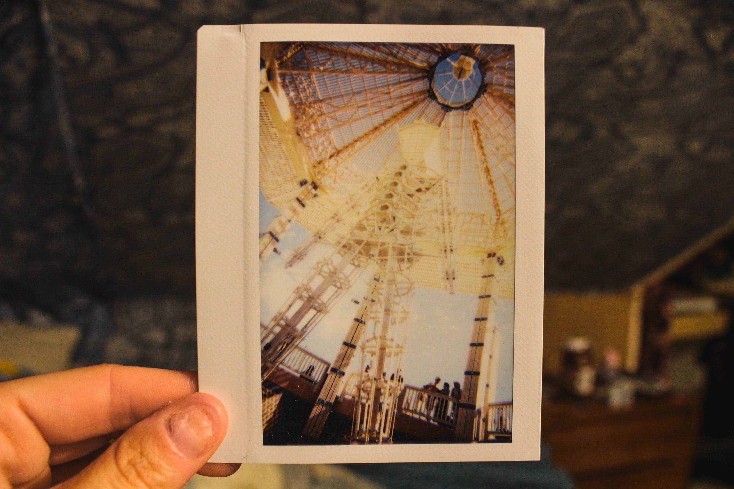 Holding a Polaroid photo of the Burning Man wooden structure as an example of what to bring to the Burn instead of small trinkets.