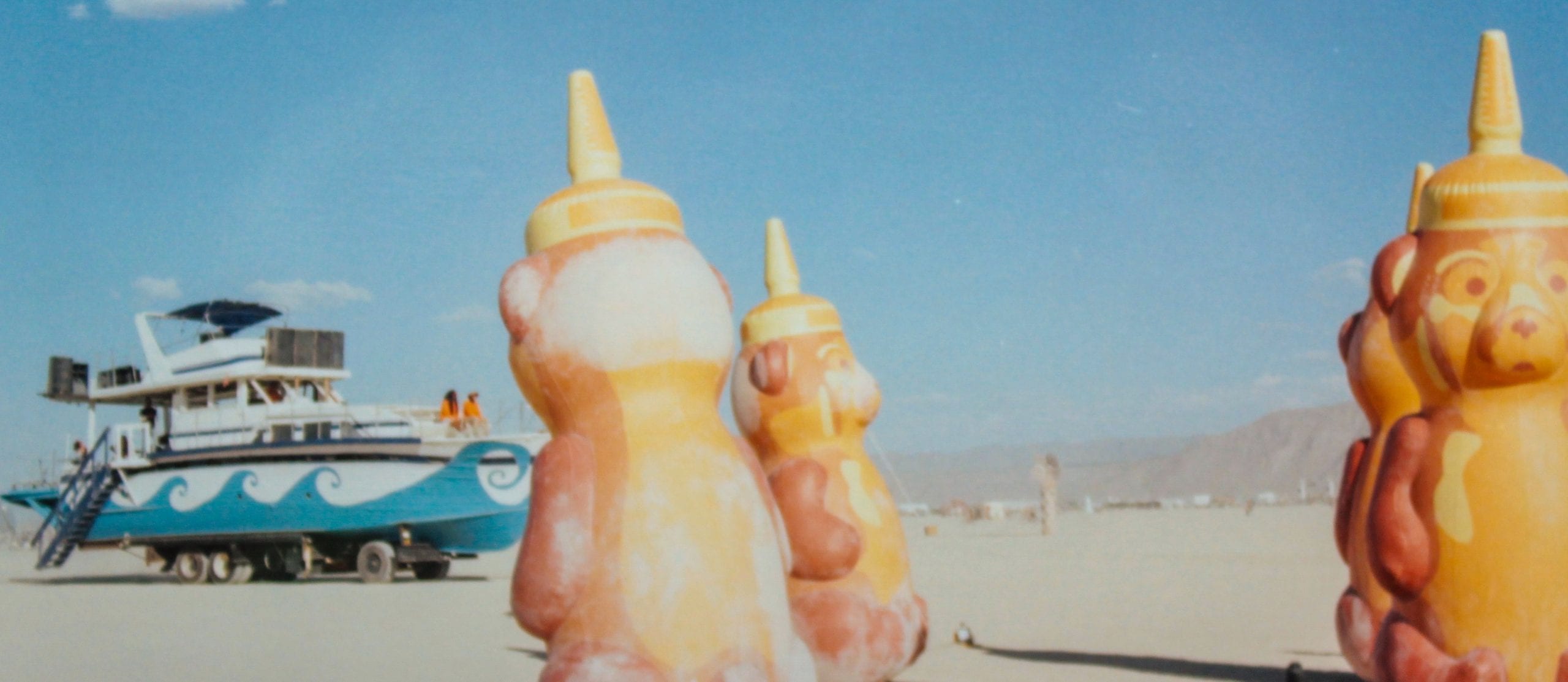 Three giant inflatable bears that resemble honey bear squeezable honey containers on the desert floor at Burning Man. In the distance is a ship art car with blue waves design on the side.