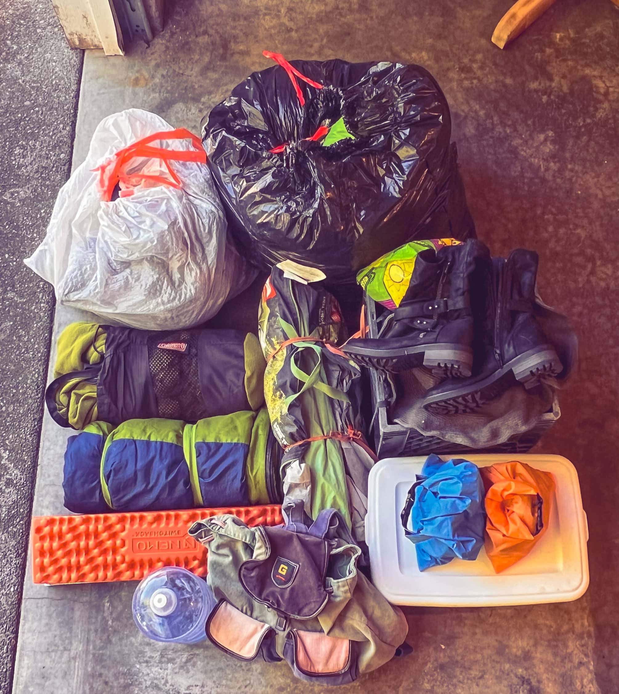 Burning Man supplies laid out on the ground including sleeping bags, pad, tent, bags of clothing, and boots.