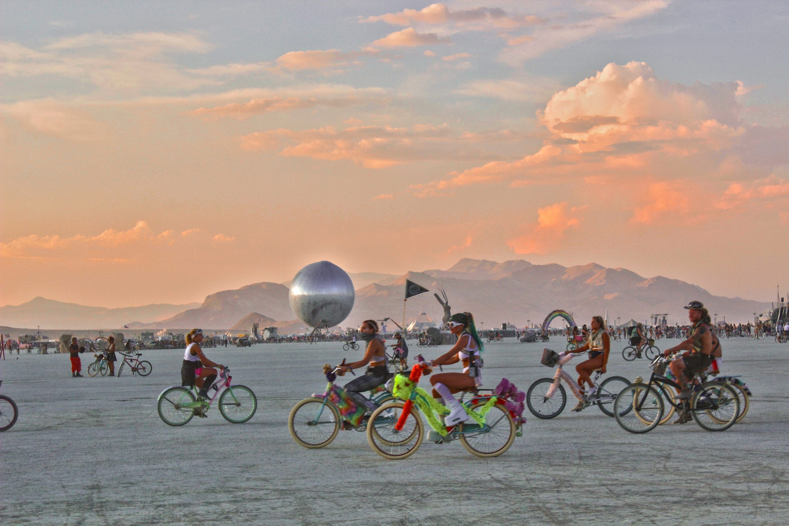 Group of bikers at Burning Man with a pink and blue sunset and the mountains in the distance. Giant reflective orb installation in distance on horizon before the hills.