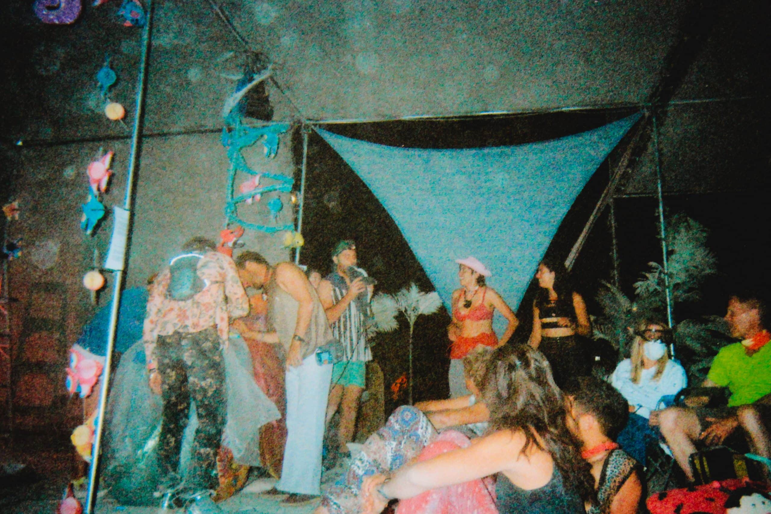 Campmates hanging out at Burning Man under a camp infrastructure at night. Fake plant decorations line the structure.