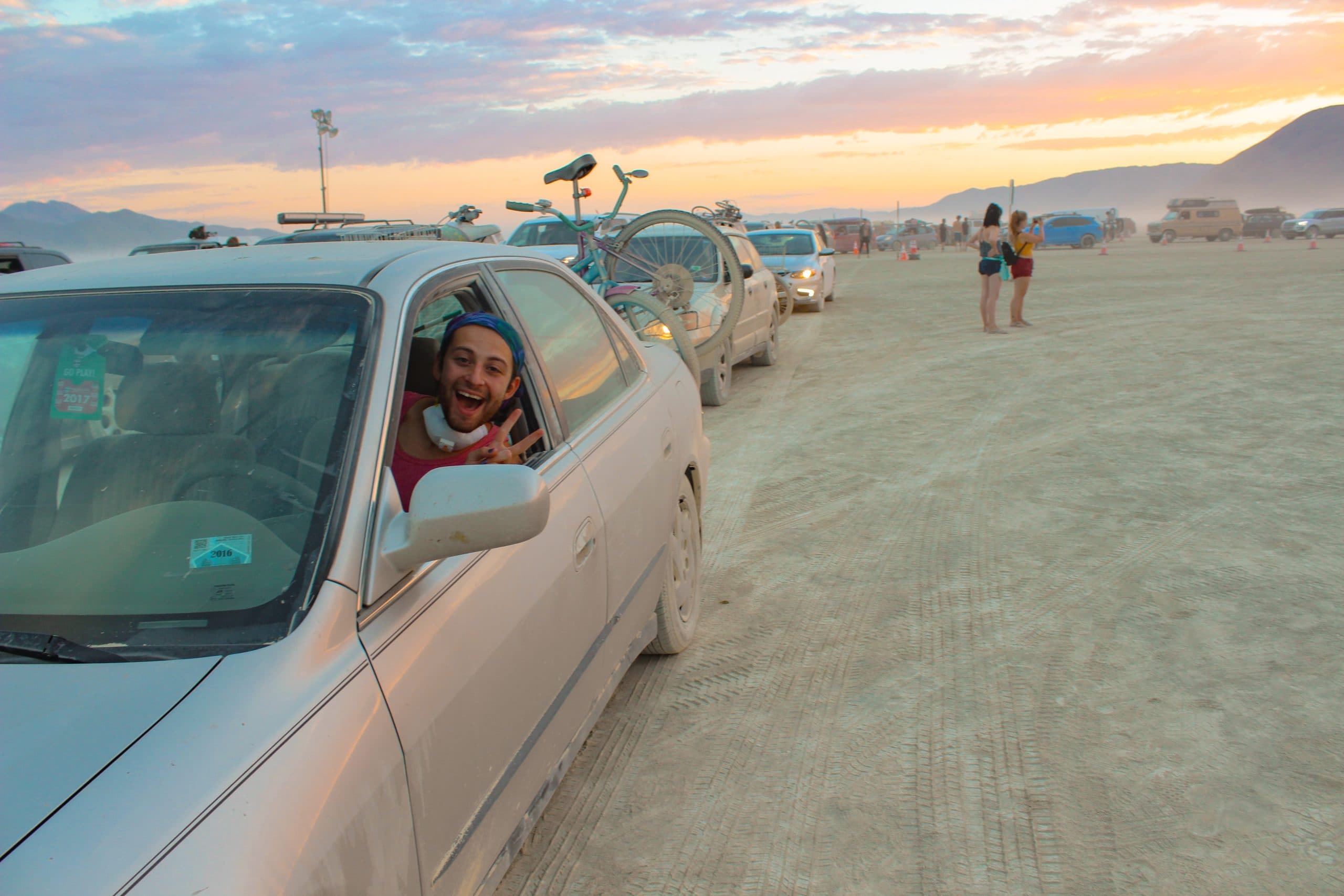 Person in a car arriving to Burning Man looking very happy. Dust on the ground and hills in the distance.