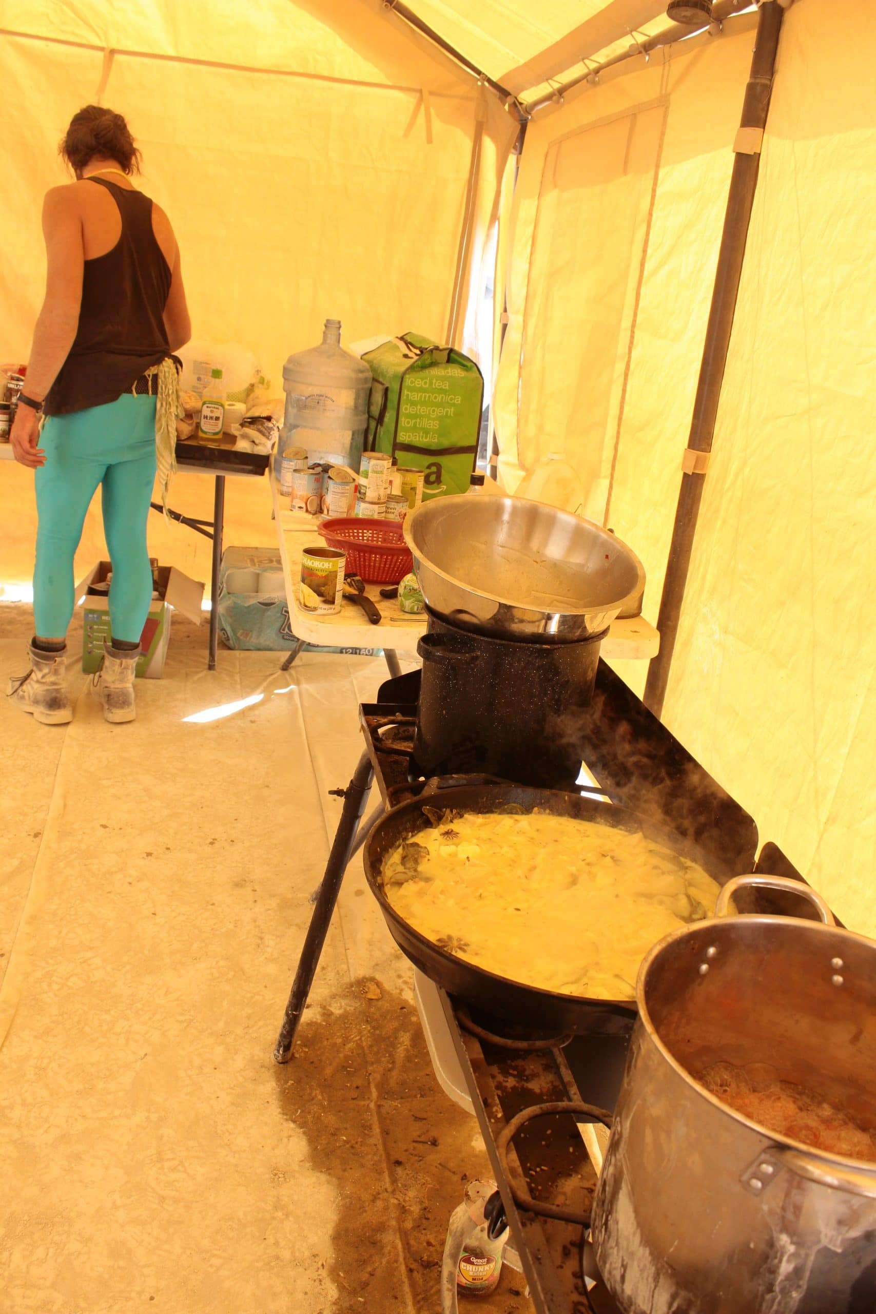 Inside a communal kitchen tent at Burning Man with a curry soup cooking on the stove top. A person next to other food and water supplies in the distance.