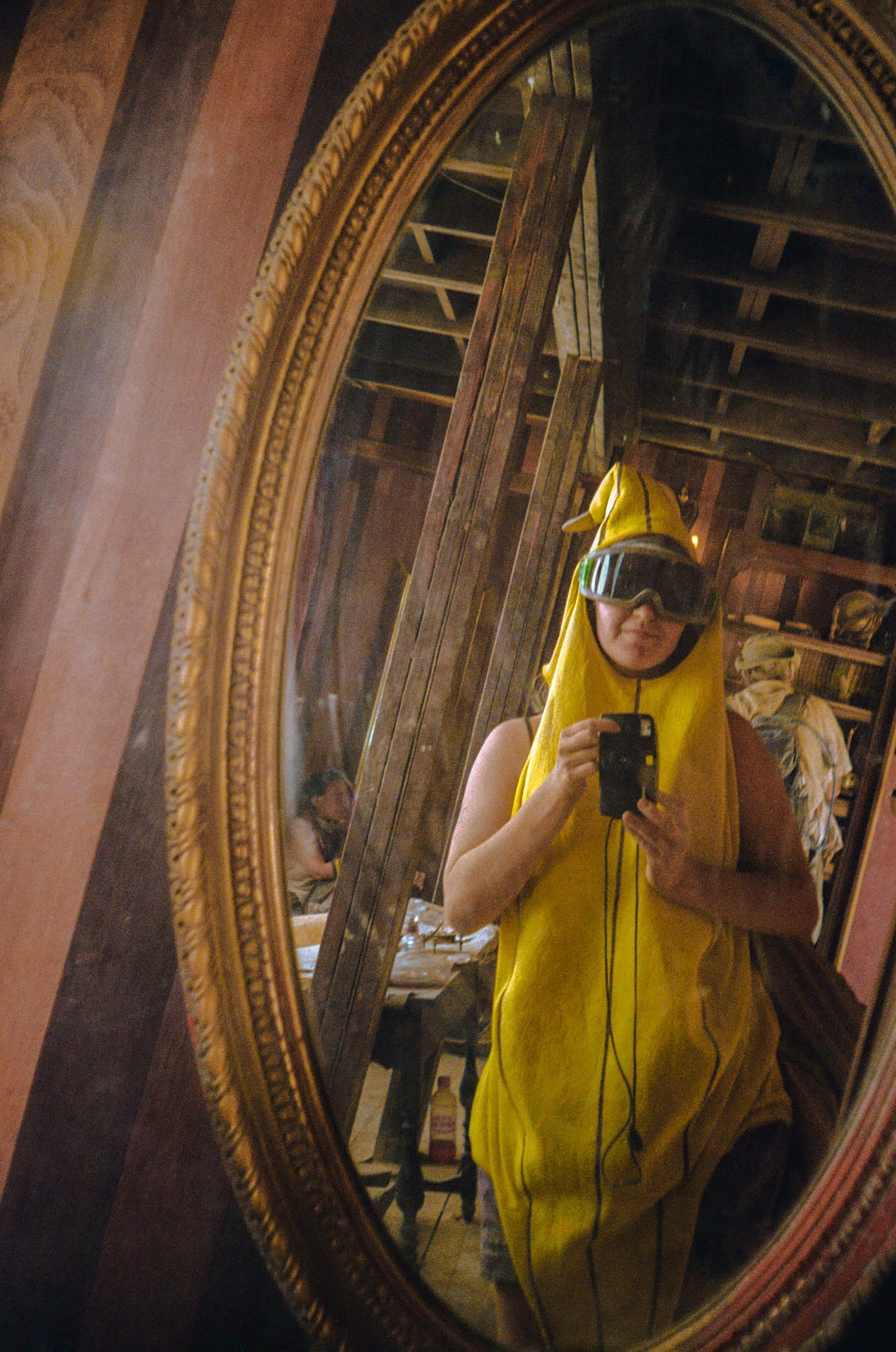 Tasha in a banana costume with dust goggles taking a photo of her reflection in an oval mirror with golden frame in a wooden style building.