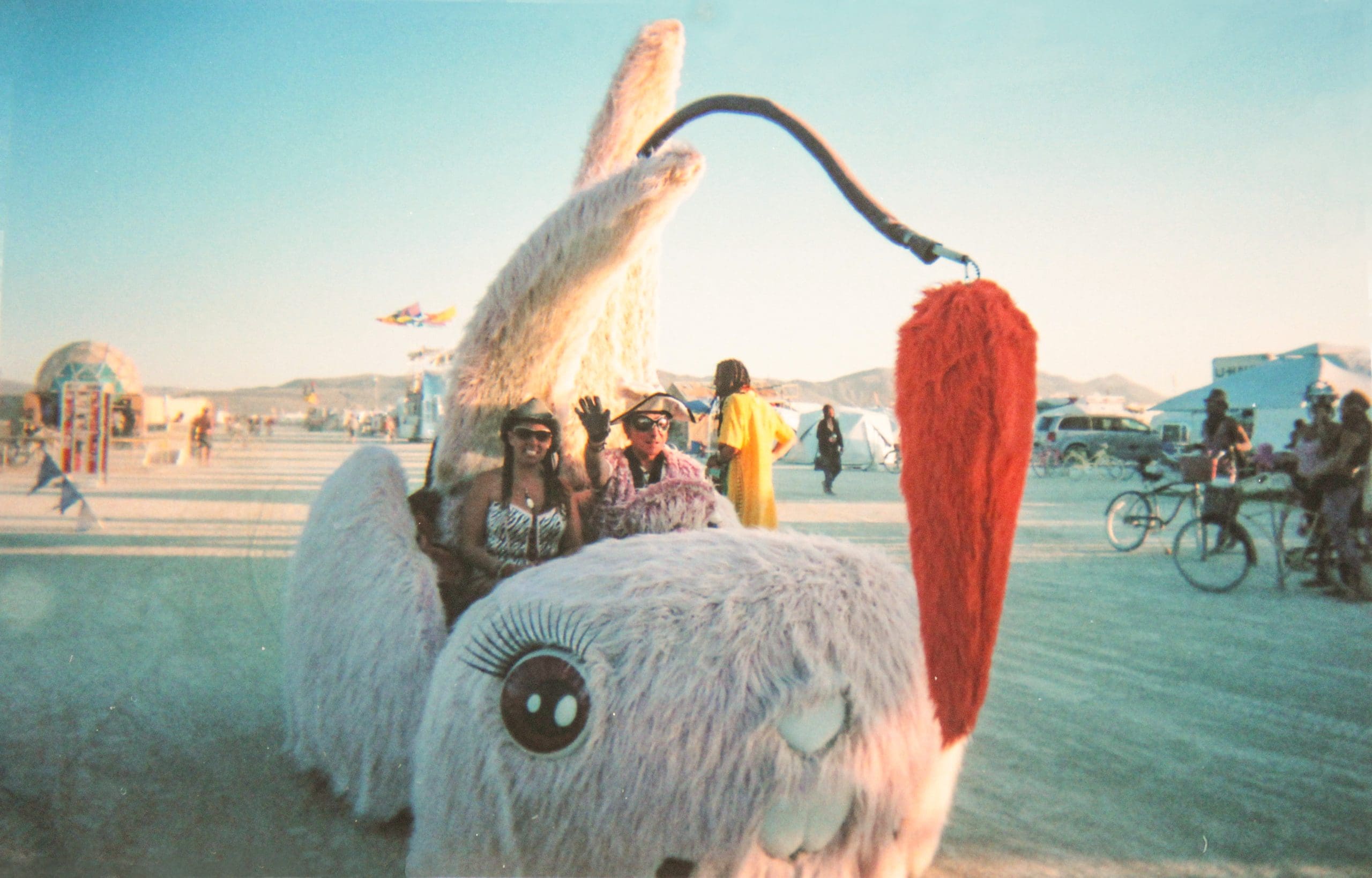 Art car at Burning Man shaped like a giant fluffy bunny with its tail coming towards the front and a hanging fake carrot. Two people are in the art car smiling.