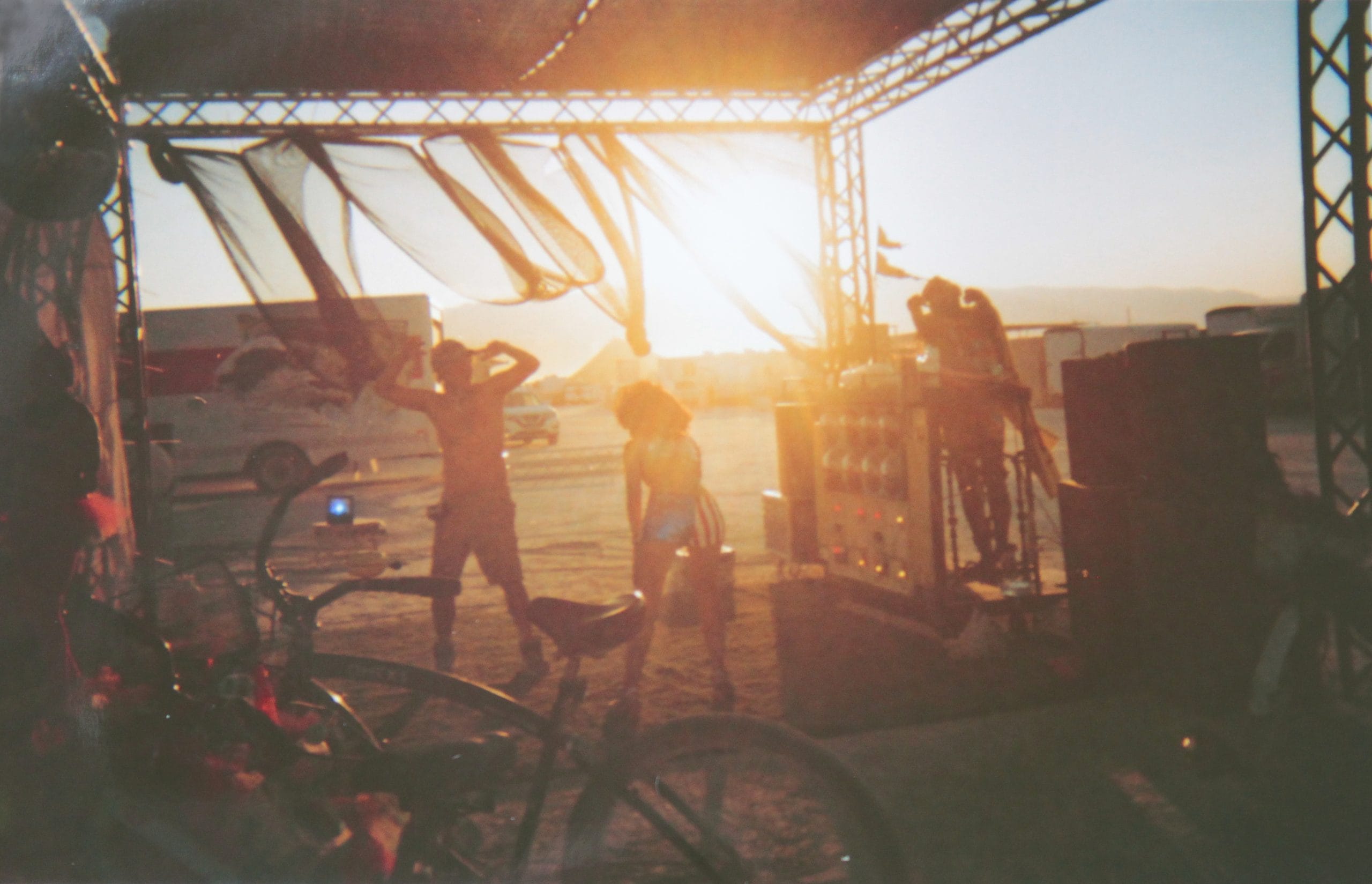 Silhouettes of two people dancing under a camp structure and the sunrise shining warm beams through in the distance.
