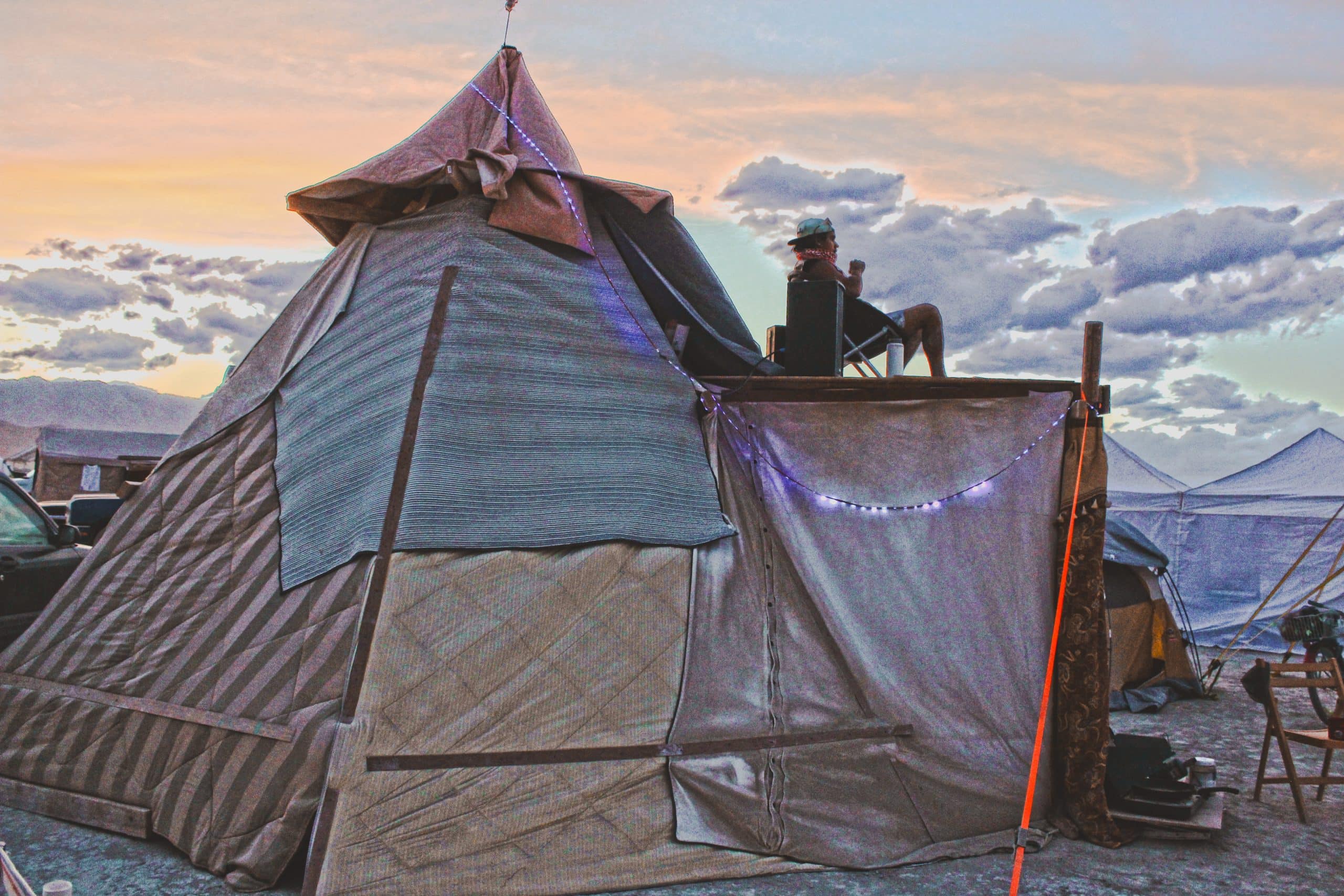 Large pyramid shaped wooden structure with blankets as walls with person sitting on the upper deck. In the distance, there are clouds and yellow sky implying sunrise time at Burning Man. Beside the pyramid there is a smaller regular Coleman tent.