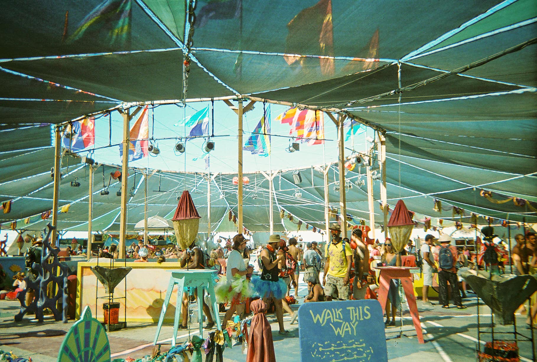 Crowds relaxing beneath the expansive shade canopy at Burning Man's Center Camp, surrounded by colorful flags, artwork, and seating areas.