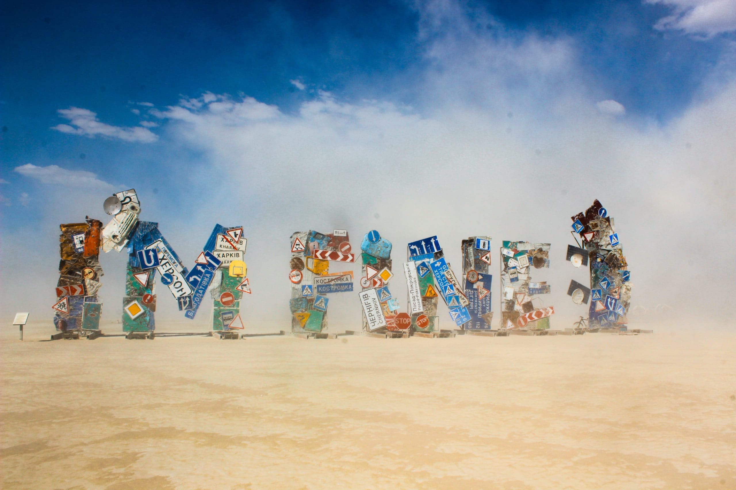 I'm Fine art installation by Ukrainian Artist at Burning Man during a slight dust storm. The art is made from street signs from the war with bullet holes in them. Blue skies and clouds above the dusty air.
