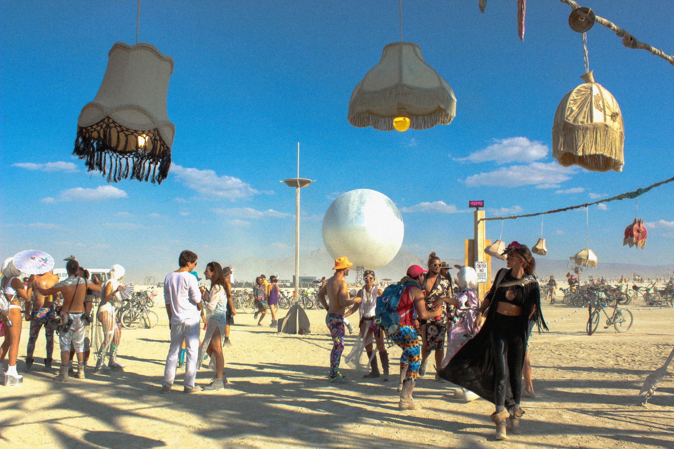 People dancing at Burning Man in the dust and sun. In the foreground three chandeliers hanging but seem to be floating. In the distance, a giant globe art installation known as the ORB --a sphere with reflective surface.