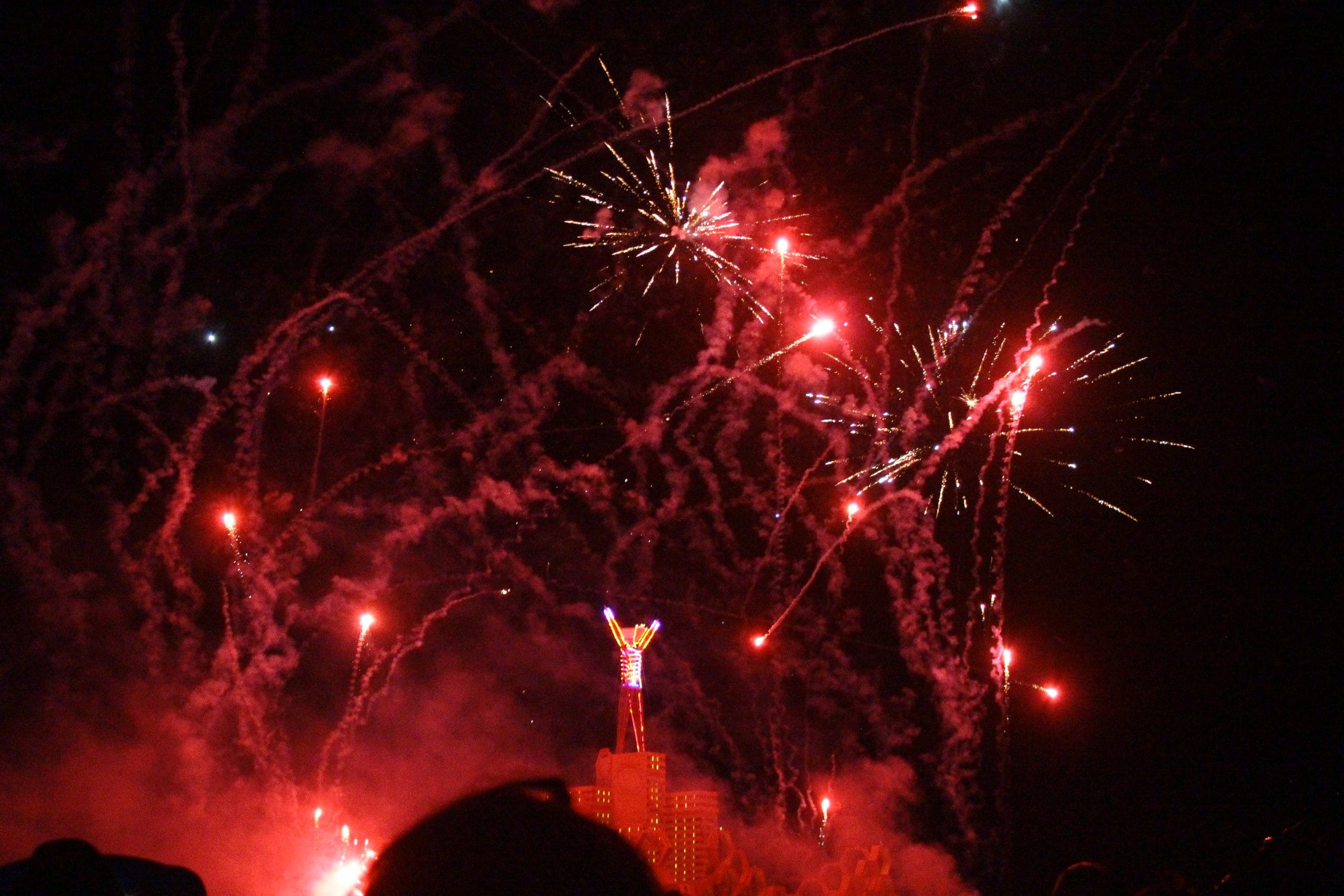 The wooden Man structure effigy at Burning Man being burned and many red firework explosions above the scene.