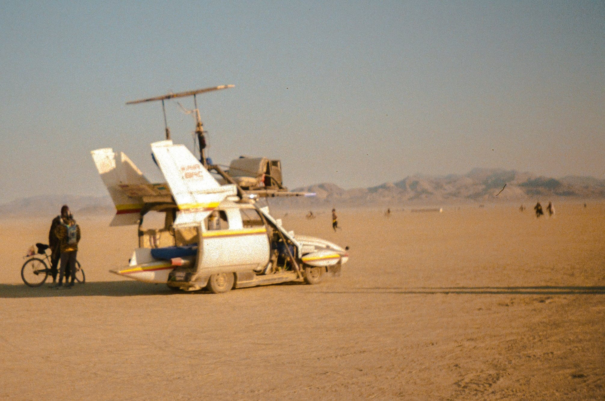 A plane art car mutant vehicle at Burning Man driving on top of the golden dust. In this distance small mountainous hills in warm glow.