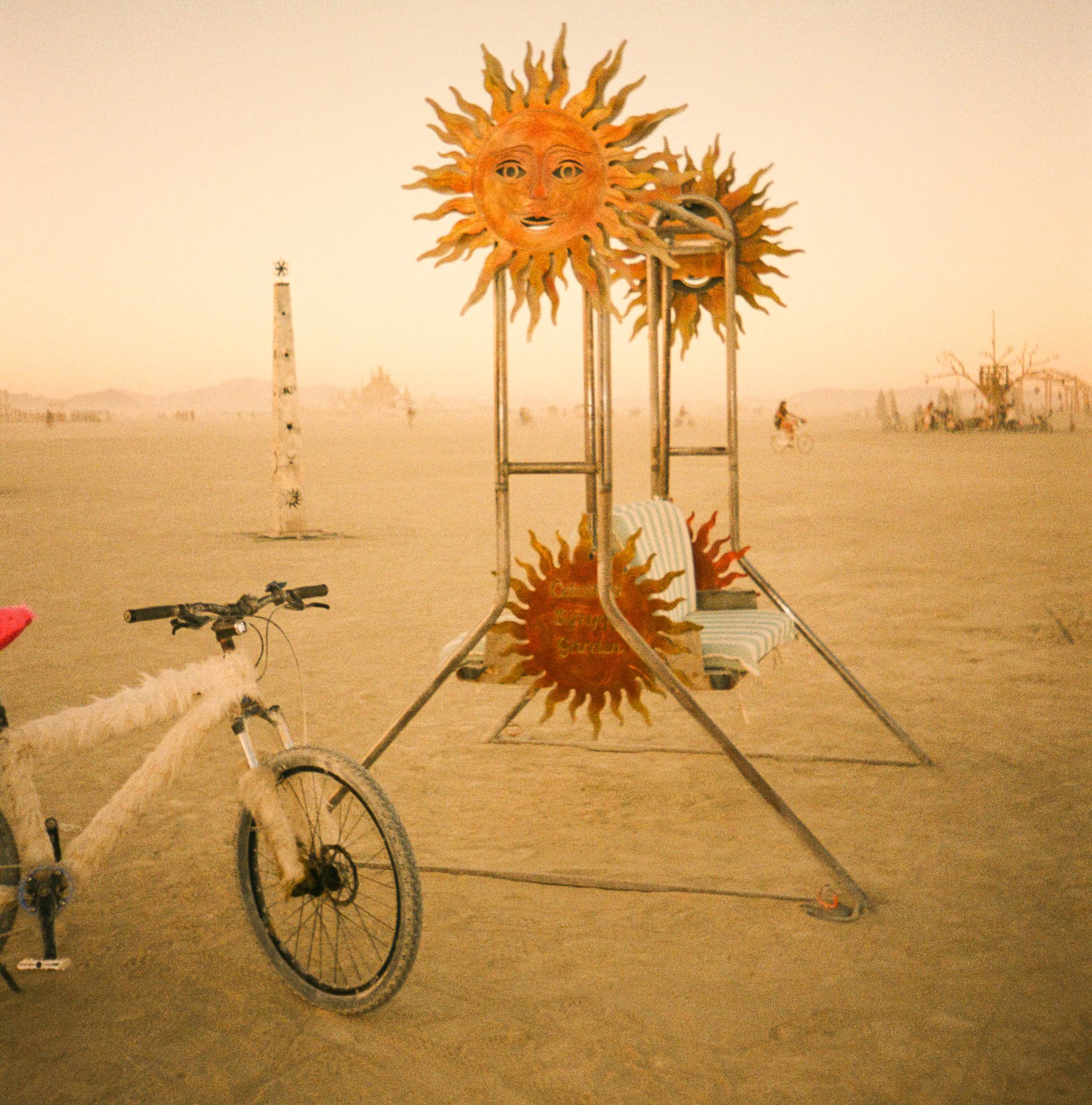 White decorated playa bike resting beside a bench adorned with smiling sun sculptures during a dusty afternoon at Burning Man.