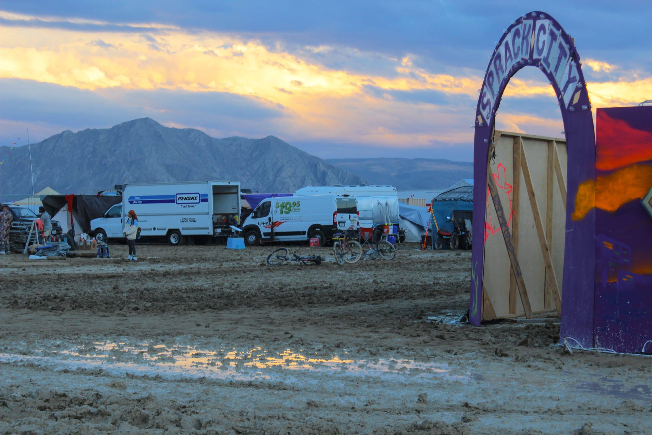Burning Man desert roads with mud and puddles showing the aftermath of rain on the playa. The hills of Black Rock Desert in the distance and yellow sunrise beams bursting through the dark rainy clouds.
