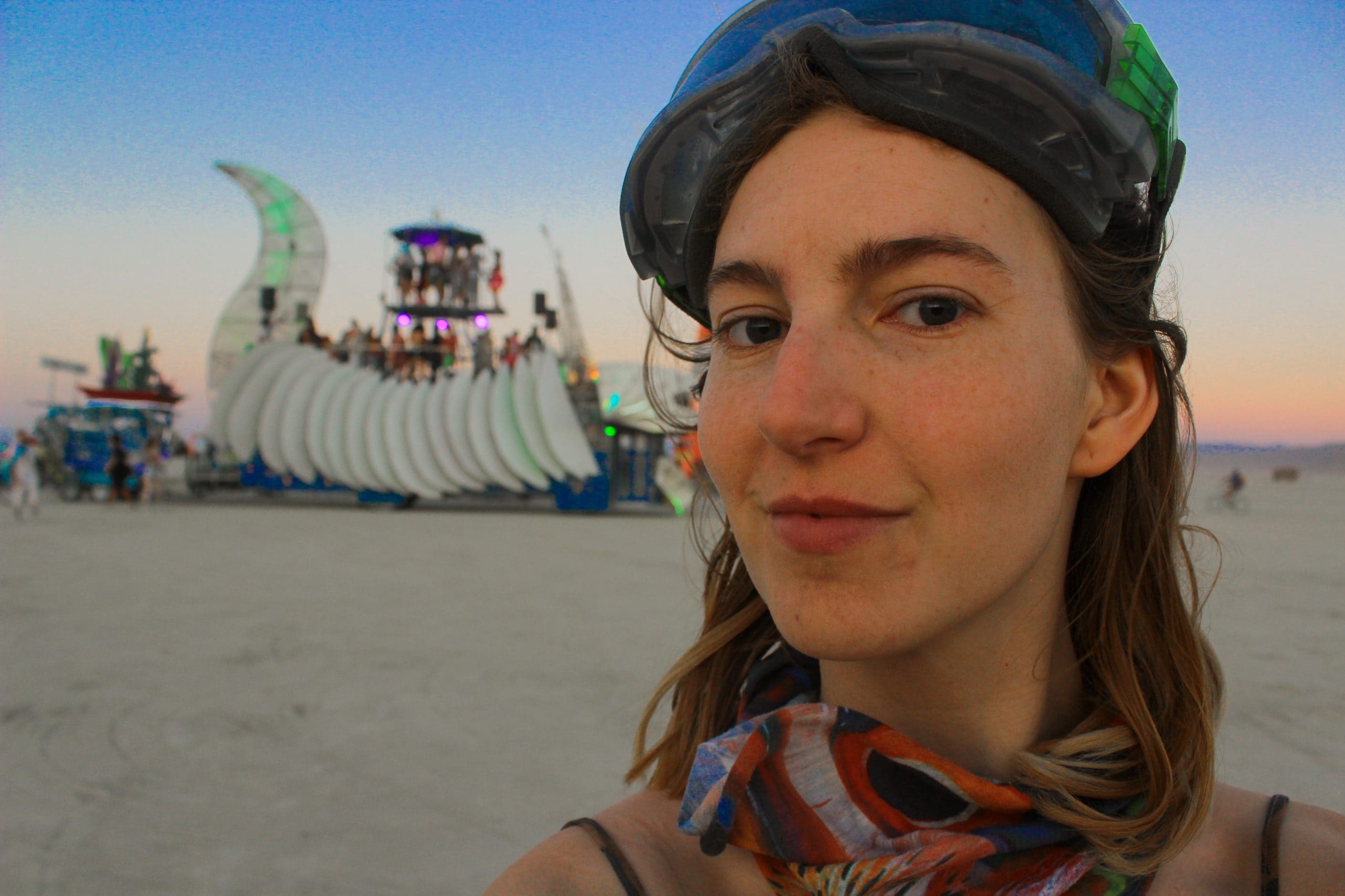 Tasha looking into the camera looking fresh and clean at the beginning of the week at Burning Man. In the distance in the sunset and an art car with people dancing on it.