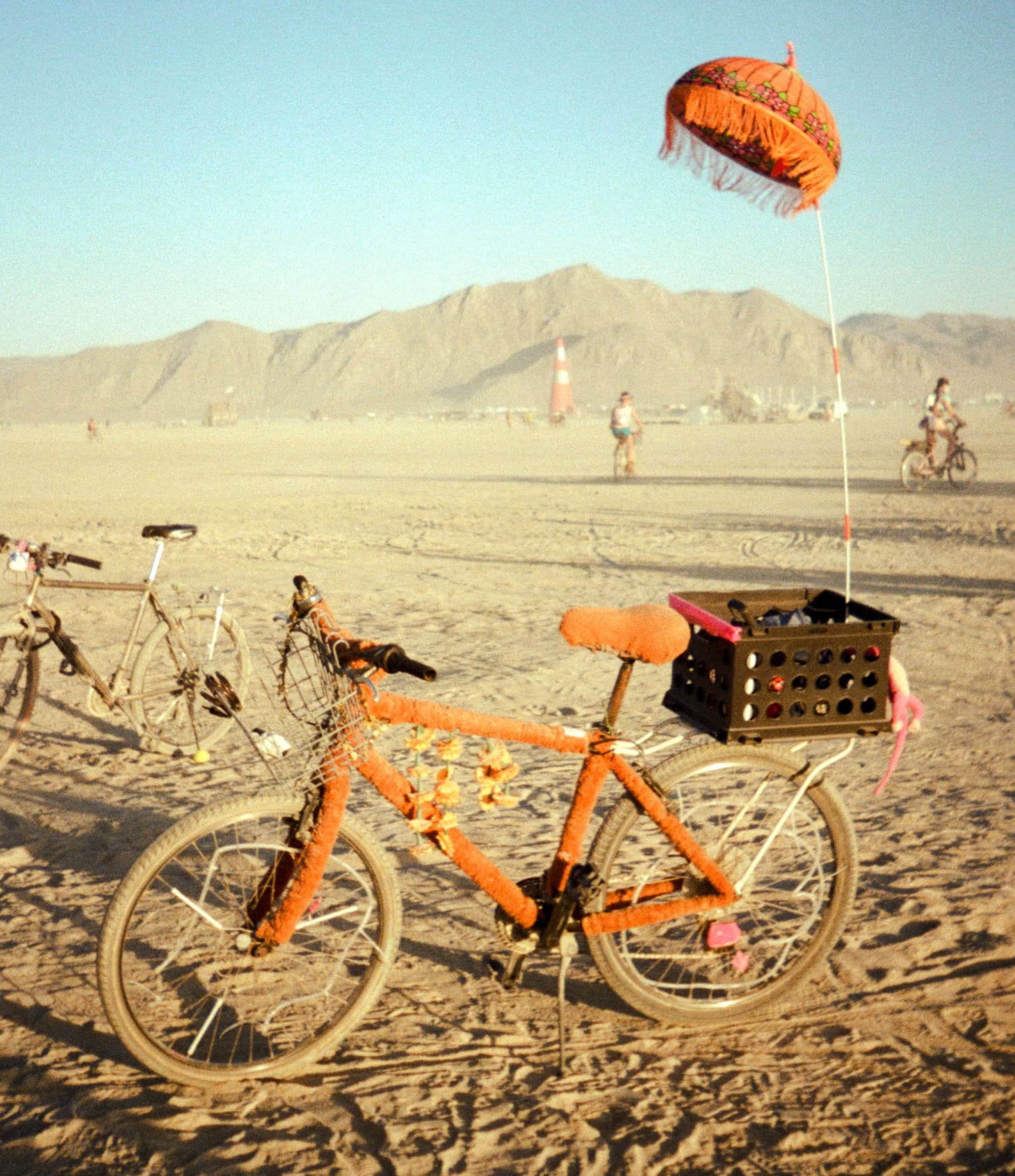 A red bicycle with a milk crate as a bike basket and long decoration with umbrella at the top in the desert of Burning Man with hills in the background.