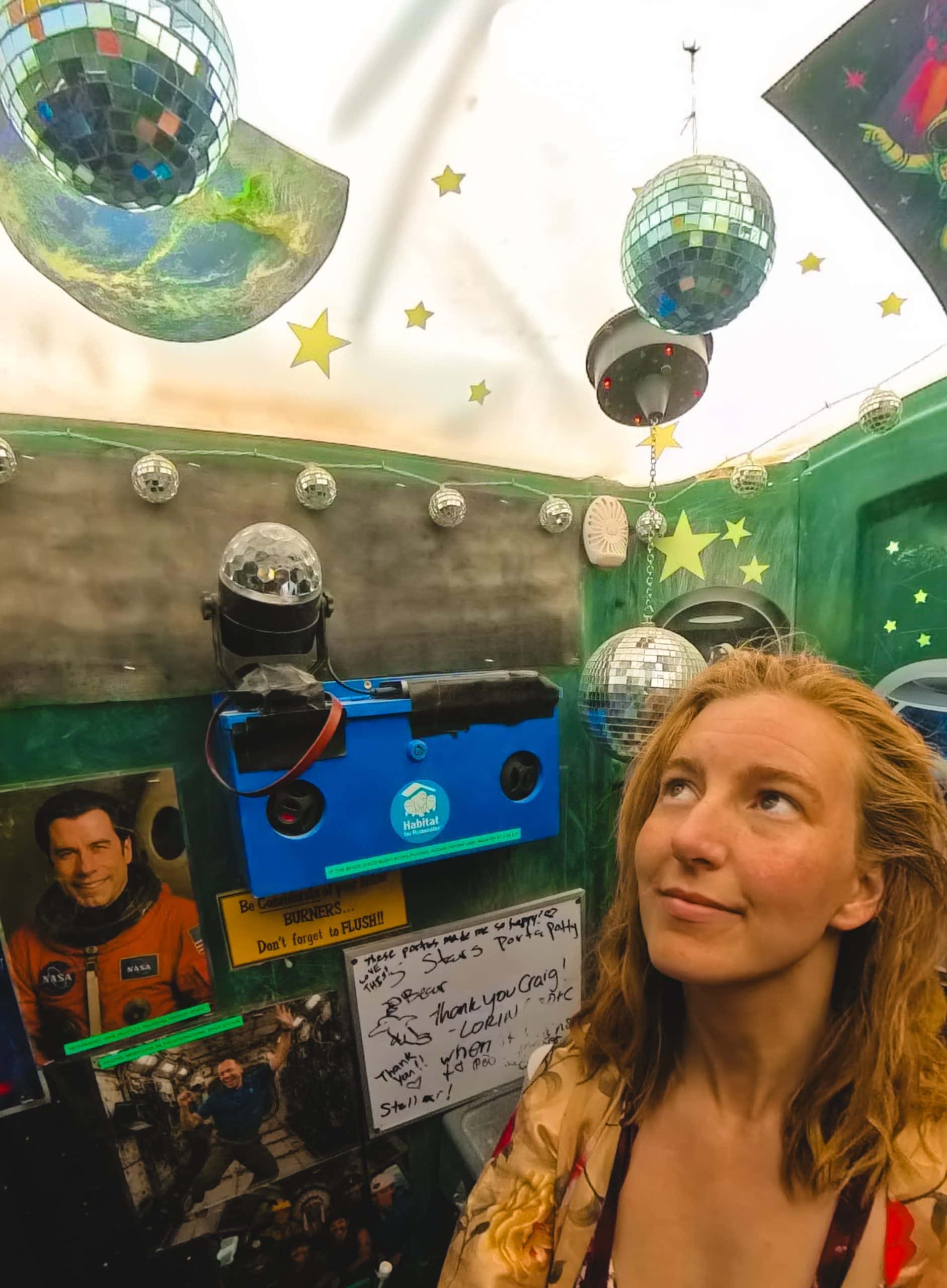 Tasha inside a porta potty at Burning Man decorated disco theme with disco balls and photos of disco singers.