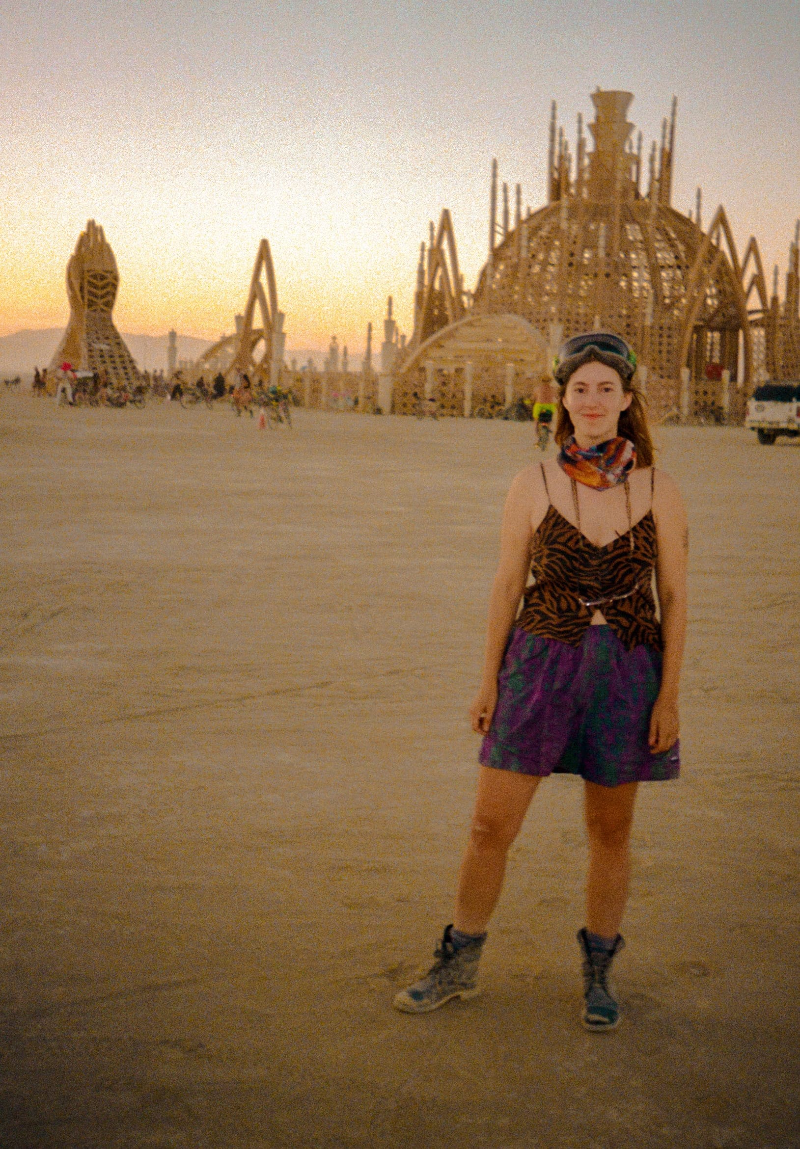 Tasha wearing stylish and practical clothing looking at the camera in the desert of Black Rock City at Burning Man. In the distance the Temple of Together from 2024--a large wooden structure with intricate design and two hands formation.