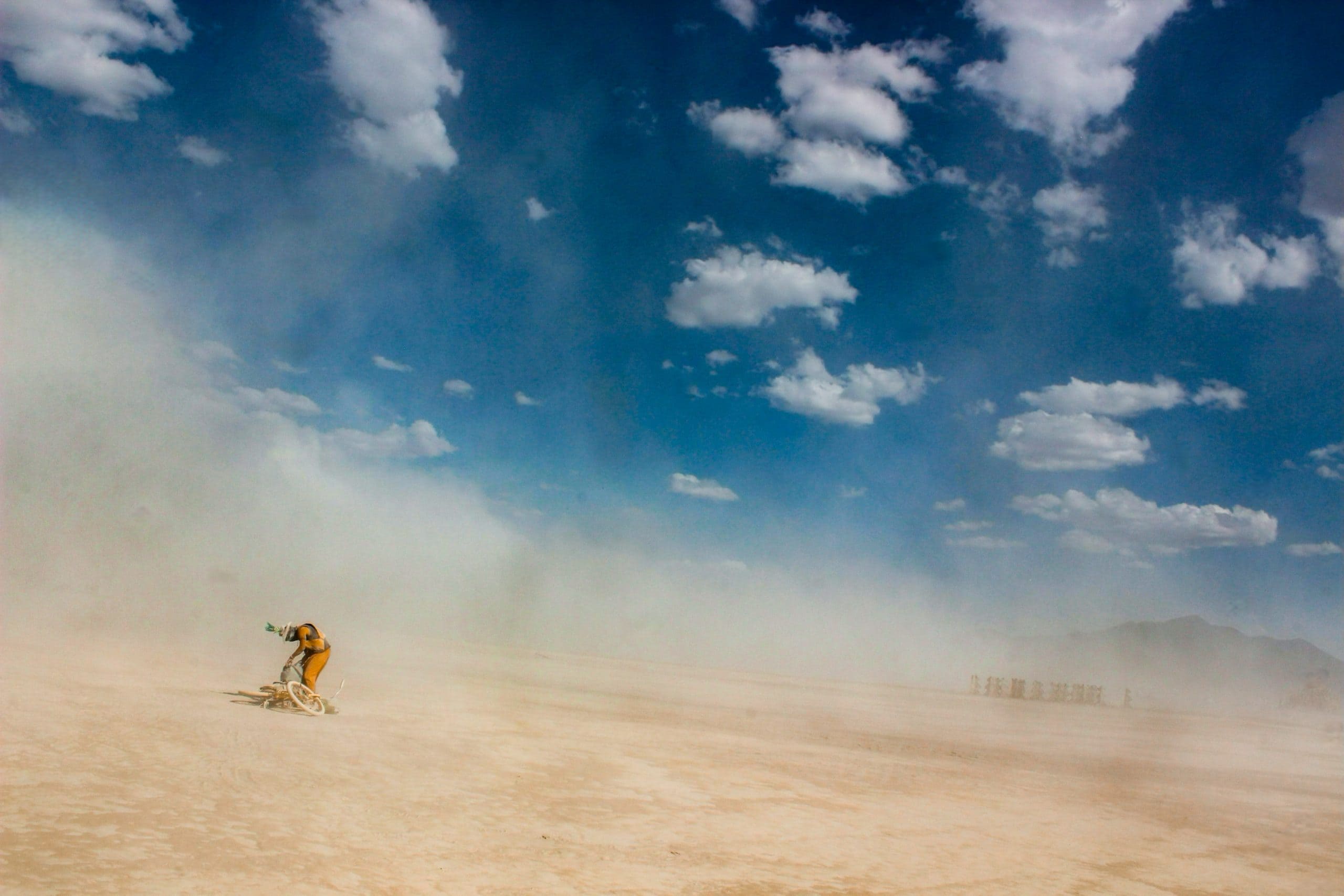 A person picking up their bike in a dust storm at Burning Man with deep blue skies and some clouds. Surrounding desert hills in the distance.