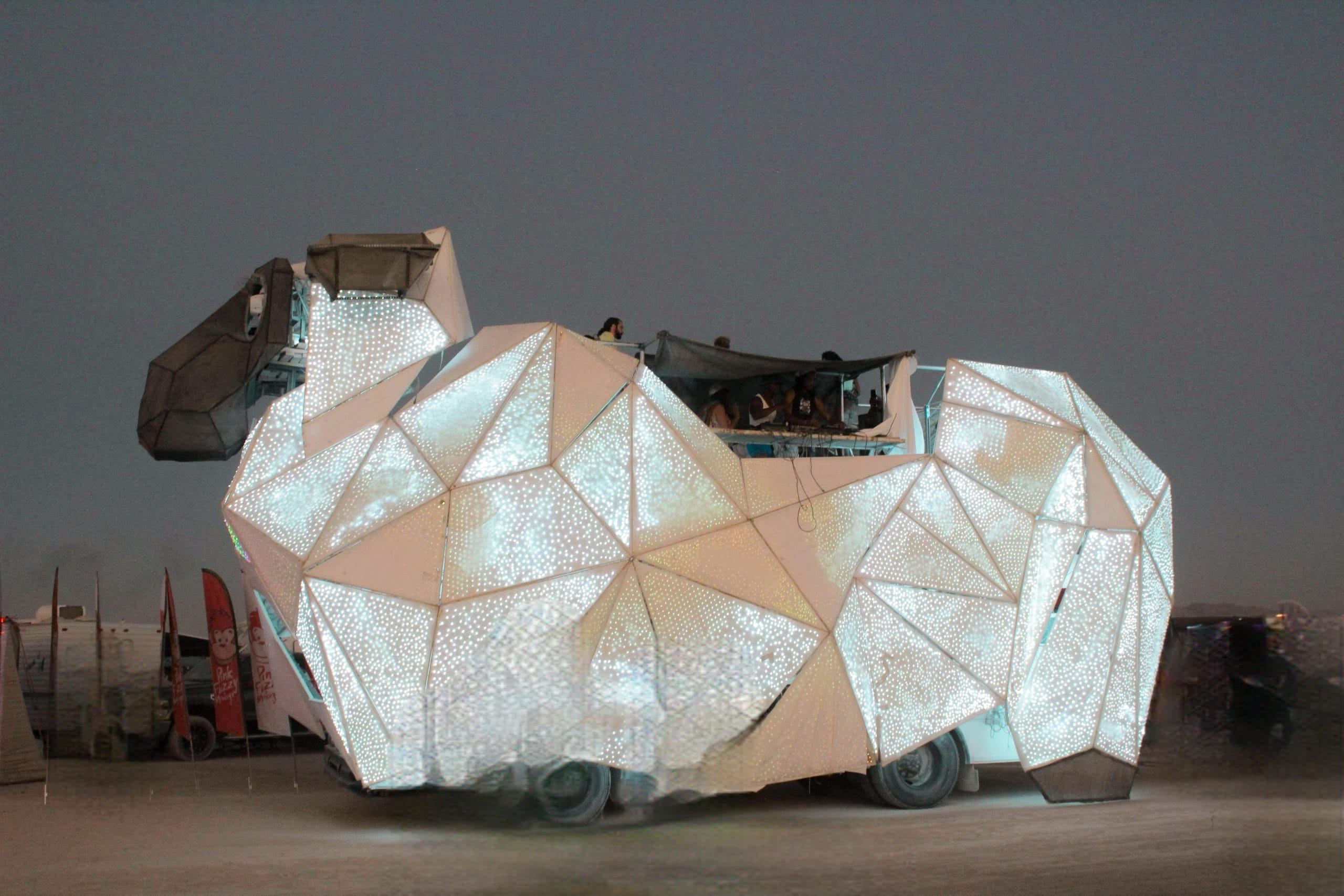 Large art car at Burning Man in the desert shaped like a sheep at dusk. The sheep fleece is made from fiberglass sheets and are illuminated so the sheep called BAAAHS  is glowing.