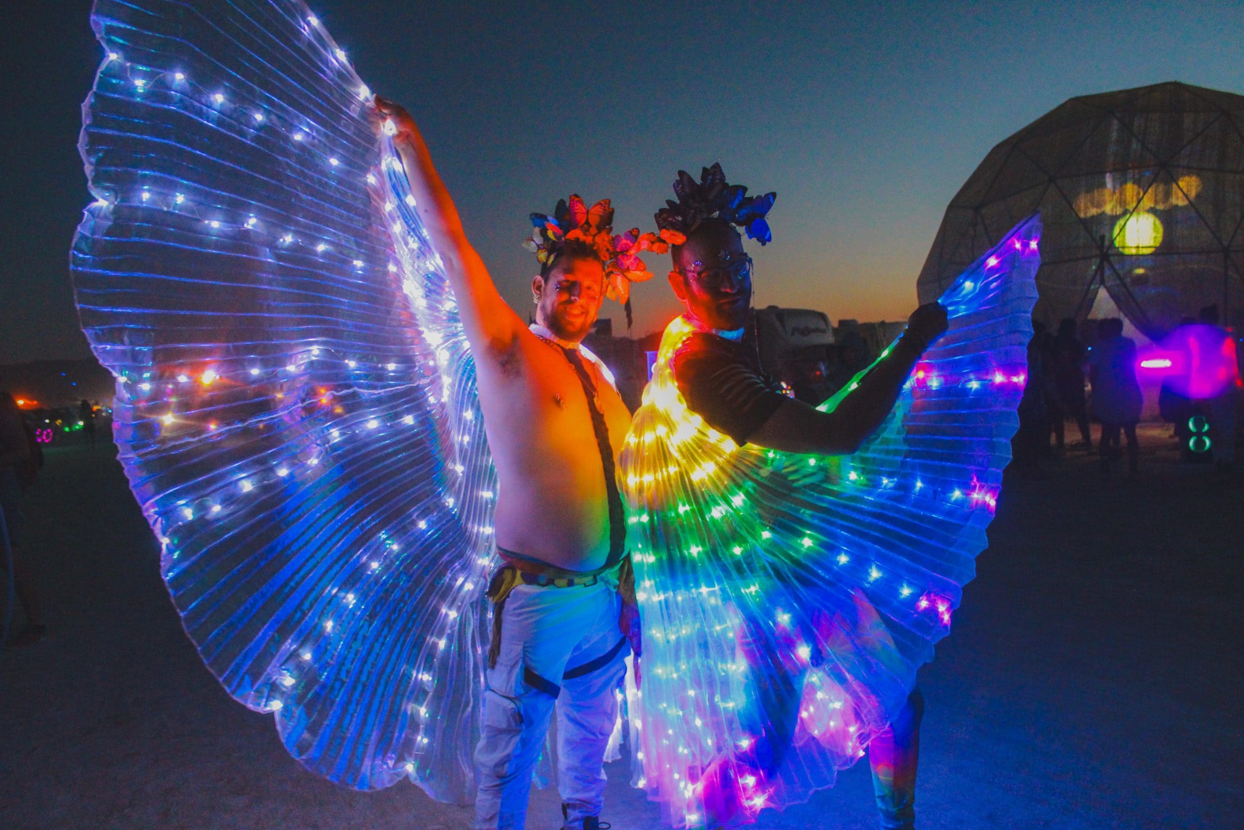 Two lovers at Burning Man at night posing with their vibrant lit up rainbow LED capes.