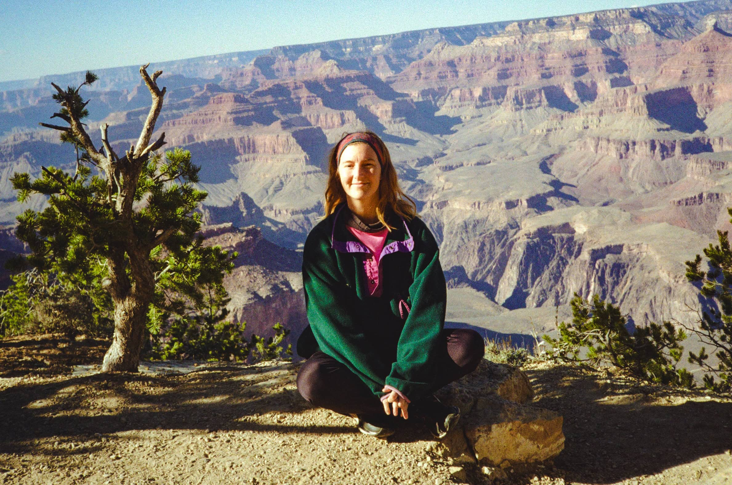 Tasha looking into the camera and sitting down on a ledge, and in the distance the hills of the Grand Canyon with deep purple shadows.
