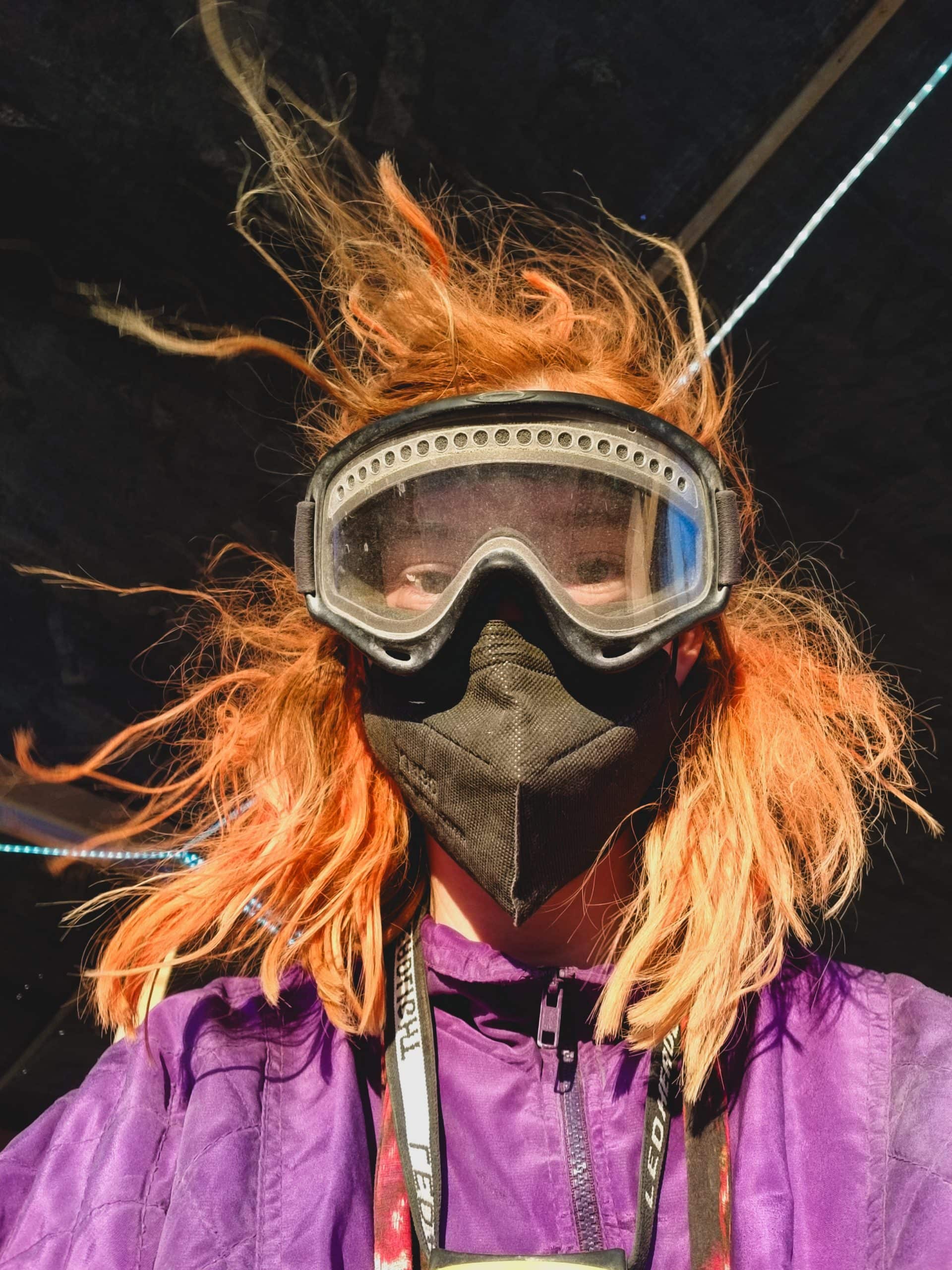 Tasha looking into the camera with wind in her dusty red hair, wearing dust goggles and a face mask for her volunteer shift.