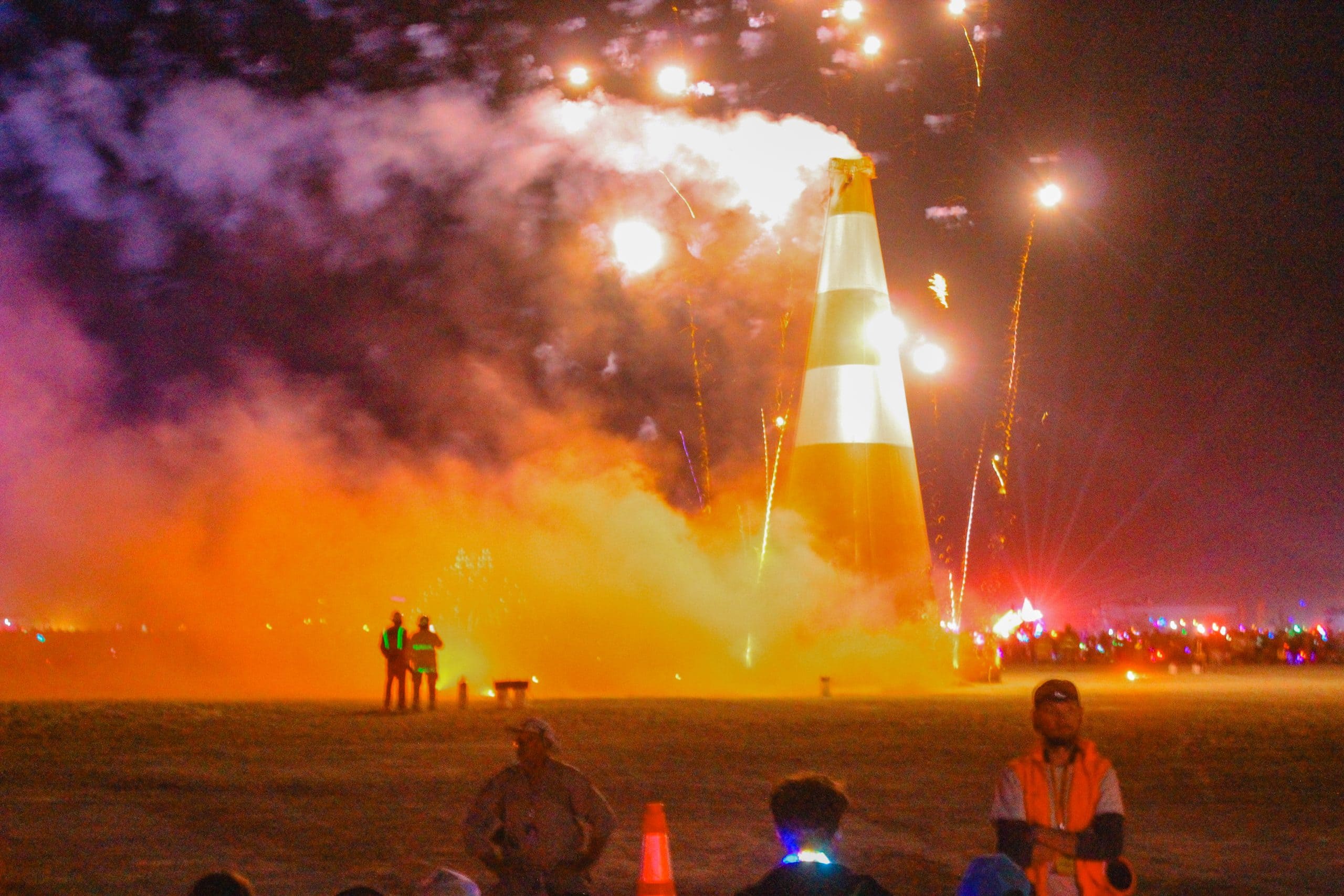 Giant traffic cone art installation called Coney McConeface burning with fireworks and orange smoke. A few volunteer staff around the burn perimeter.