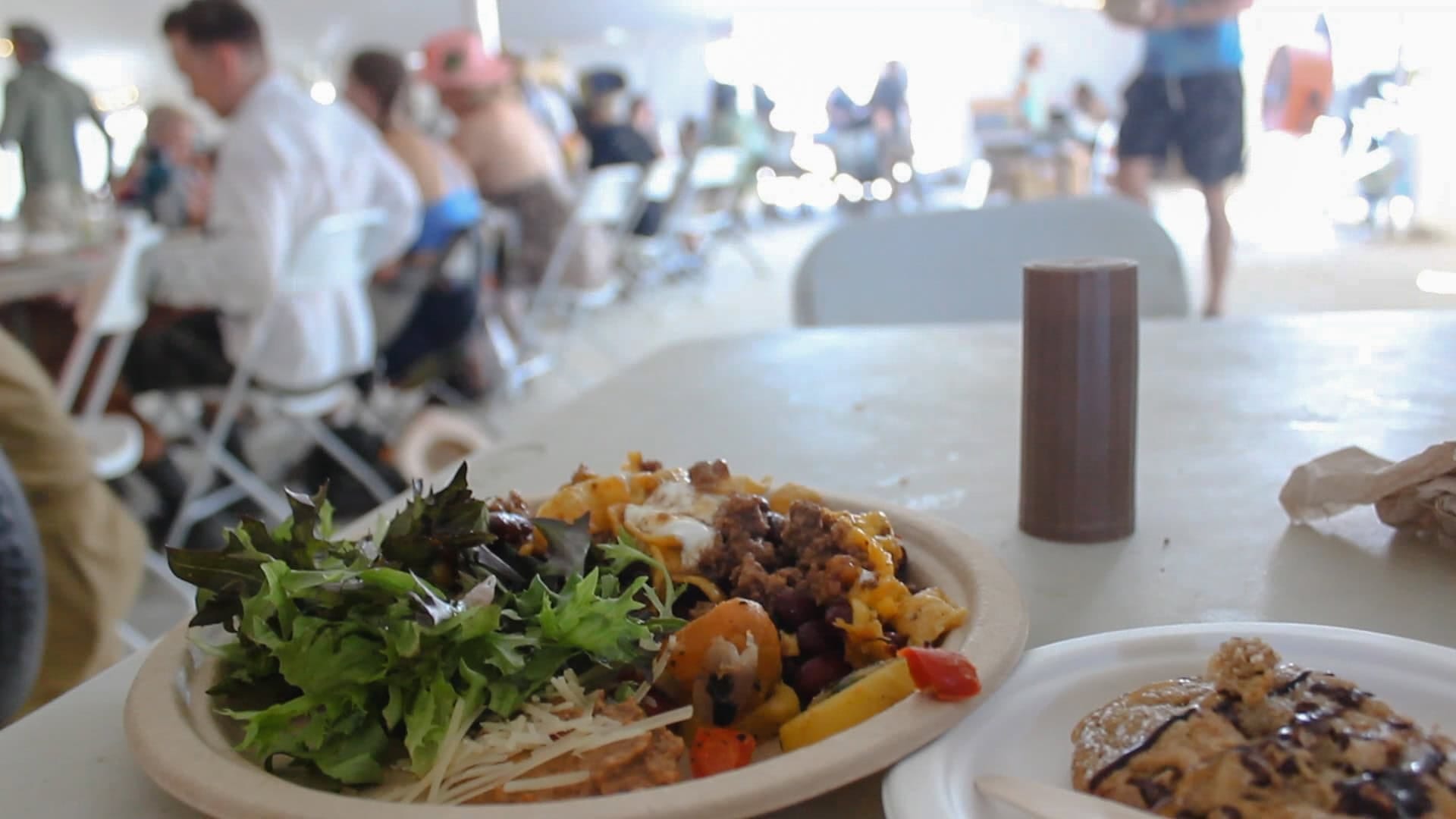 Two plates of food with salad and a cookie on a table. Background is slightly out of focus sight of people sitting down eating in a large dome at Burning Man.