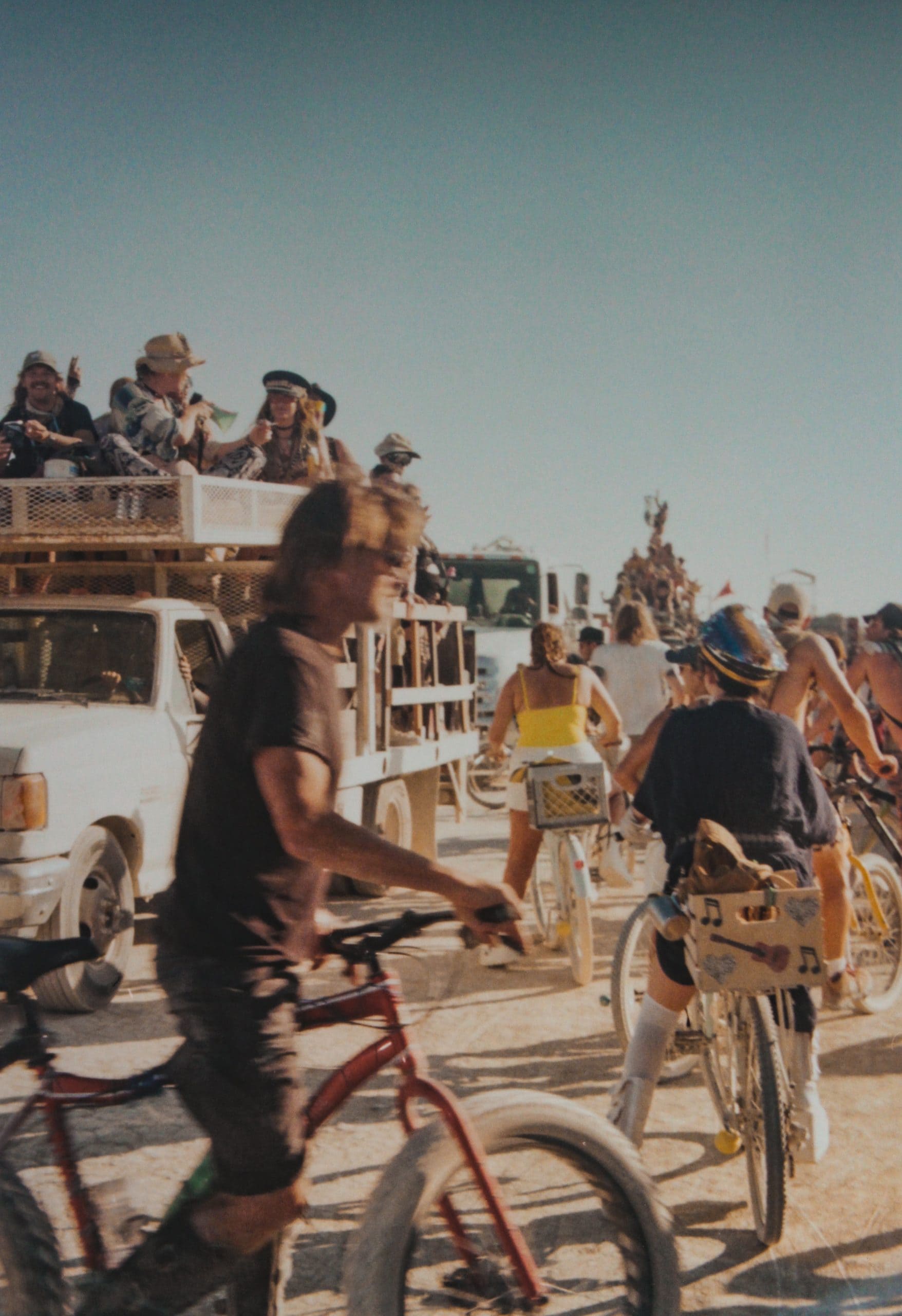 The GP&E annual parade at Burning Man with a line of cars and many people on top of the cars. The dusty street at Burning Man is filled with people on their bikes looking towards the car parade.