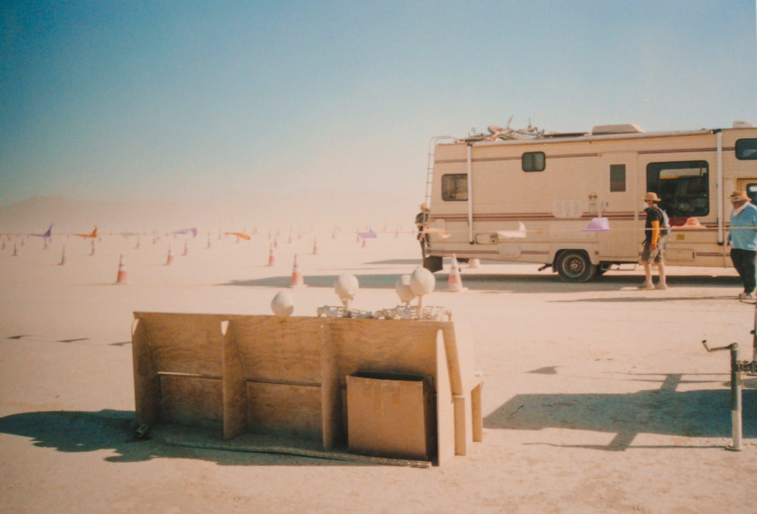 A bench with FOUR skeletons on it at the entrance Gate to Burning Man with an RV and blue skies in the distance. Driving lanes are separated by traffic cones on the dusty ground.
