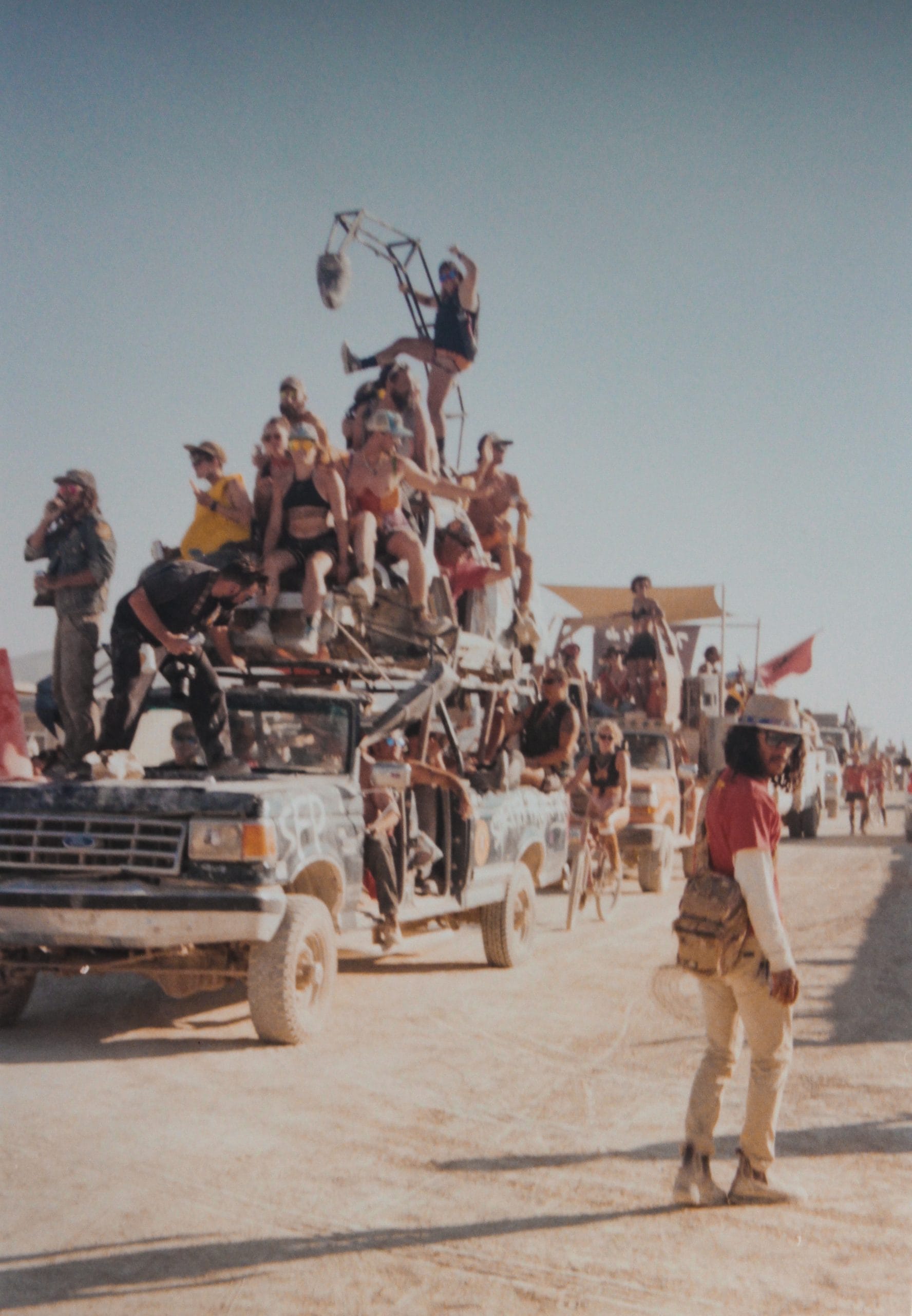 The GP&E parade at Burning Man with a line of cars with a truck with many people on top of it at the front. People are cheering and a person in front of the truck is standing on the dusty ground.