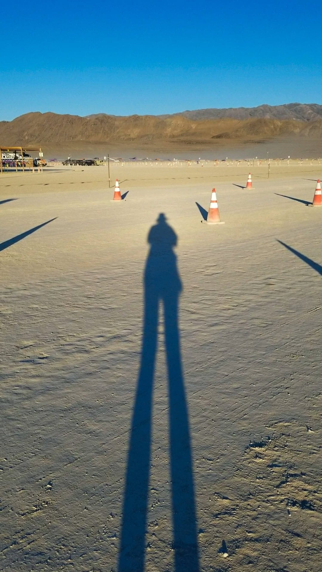 A very long shadow of Tasha from sunrise at a volunteer GP&E shift with Gate crew. Traffic cones on the dusty ground forming lines for the driving lanes although there are no cars. In this distance the hills surrounding the playa desert.