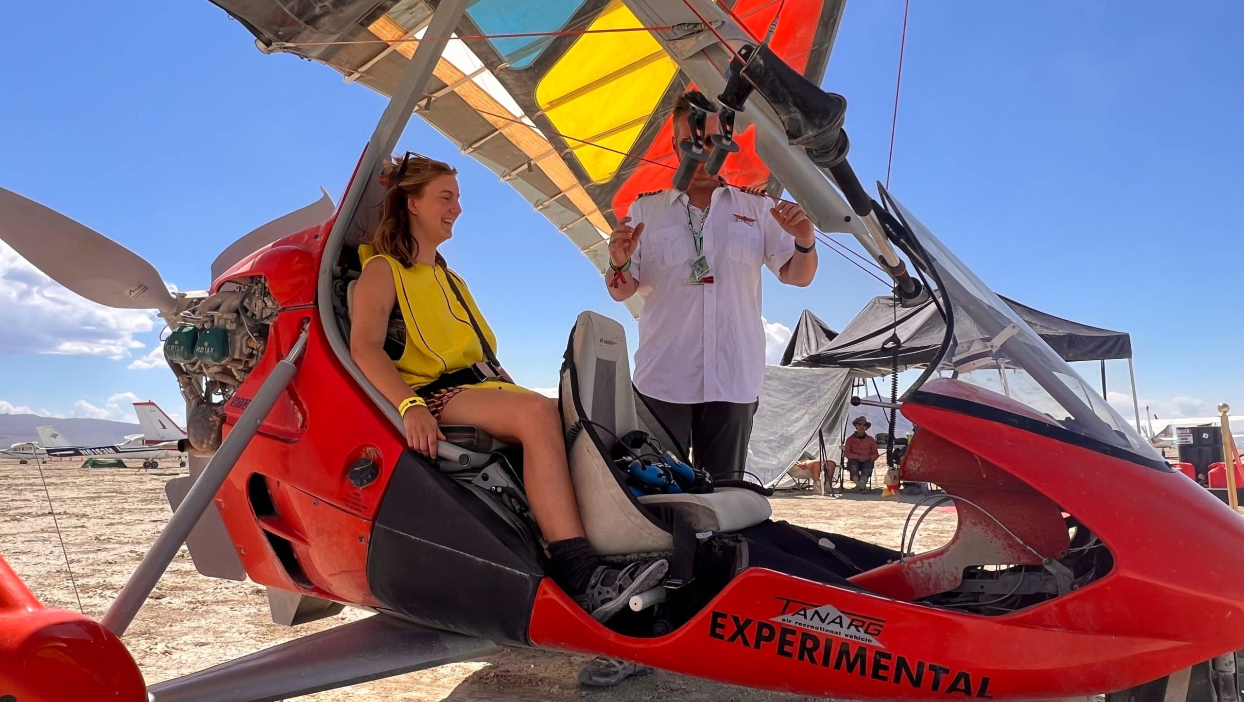 Tasha wearing a banana suit sitting in the back of a glider plane at Burning Man with the flight captain getting in his front seat. The Glider plane is bright red with a yellow and red vibrant kite above.